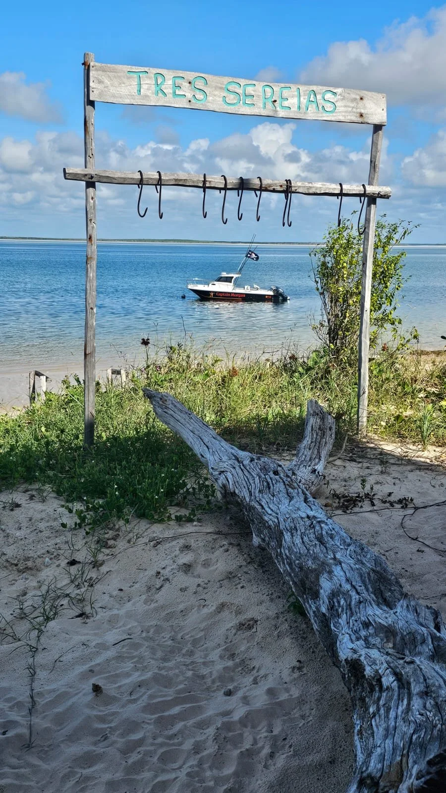 Beach scene with a wooden sign reading 'Tres Serias' and several nooses hanging from it, a small boat anchored in the water, sandy shore with driftwood and greenery, under a partly cloudy sky.