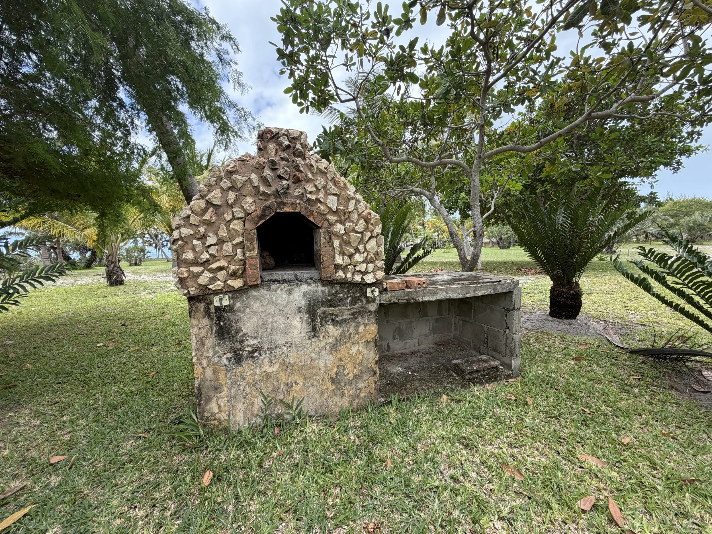 Stone outdoor fireplace with chimney, set in a grassy park area under trees with lush green foliage.