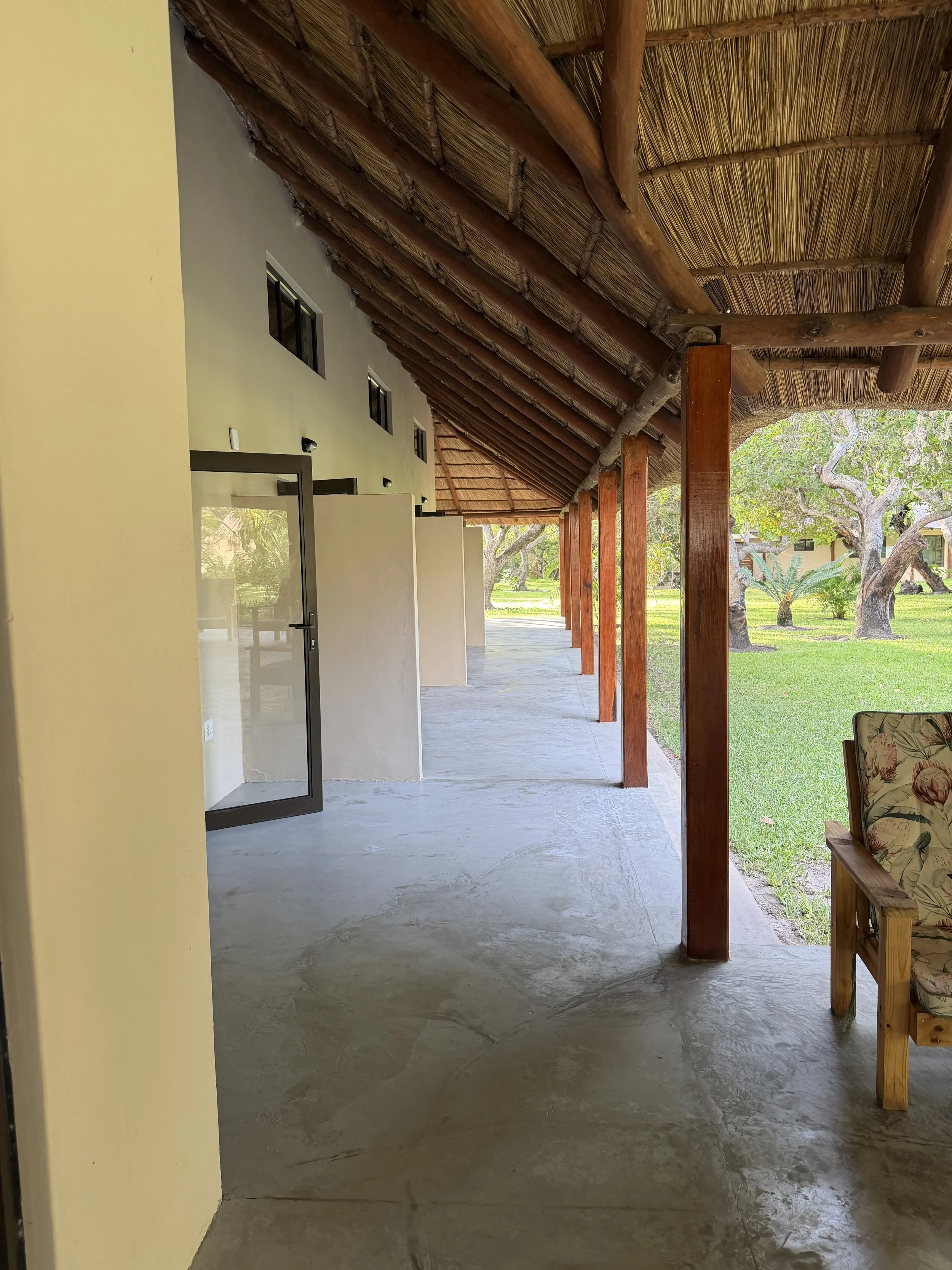 View of an outdoor covered patio area with a thatched roof, wooden support beams, a concrete floor, and a chair with a floral cushion. In the background, there is green grass and trees.