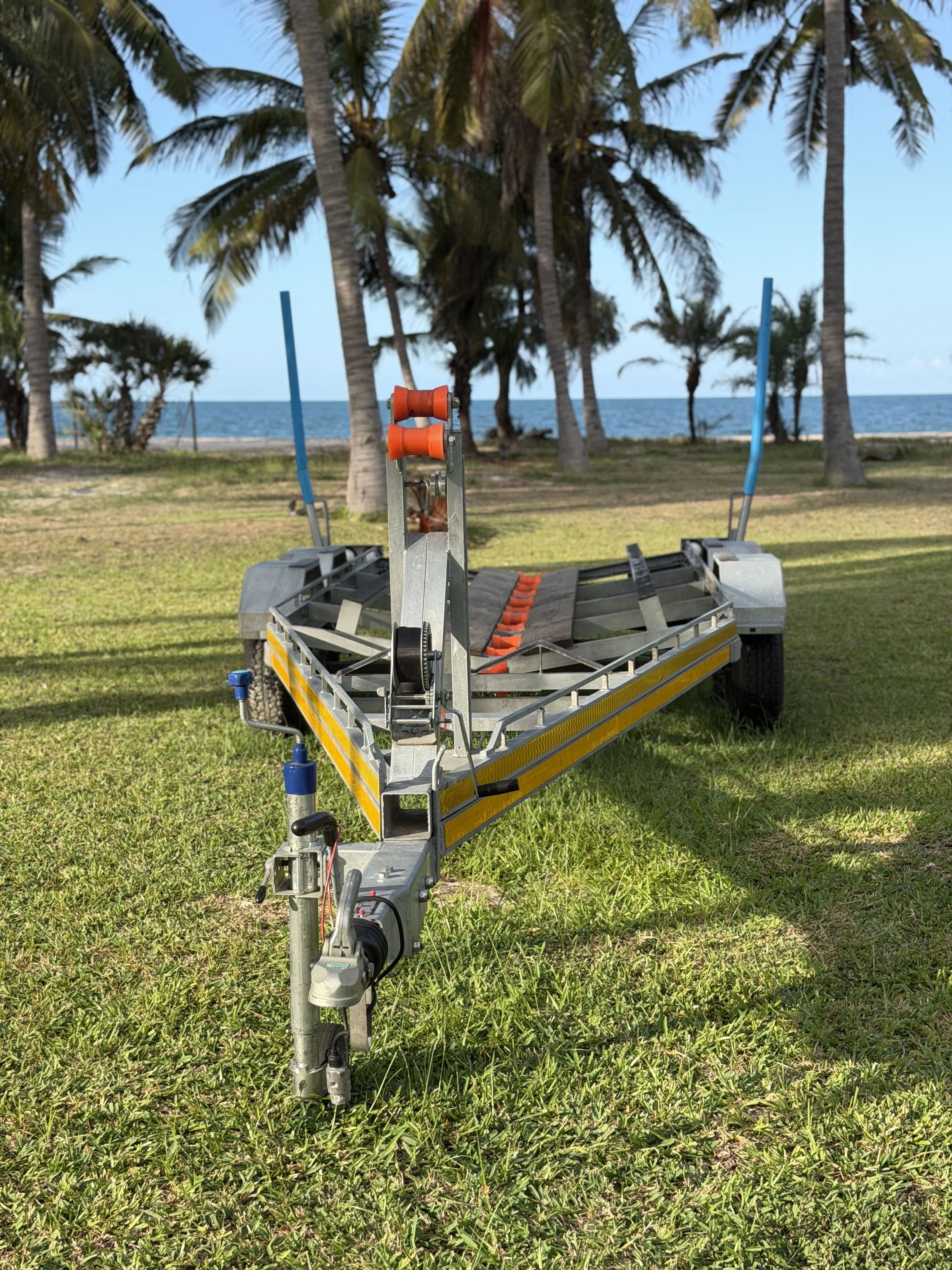 A boat trailers on green grass near the beach with tall palm trees in the background and the ocean beyond.