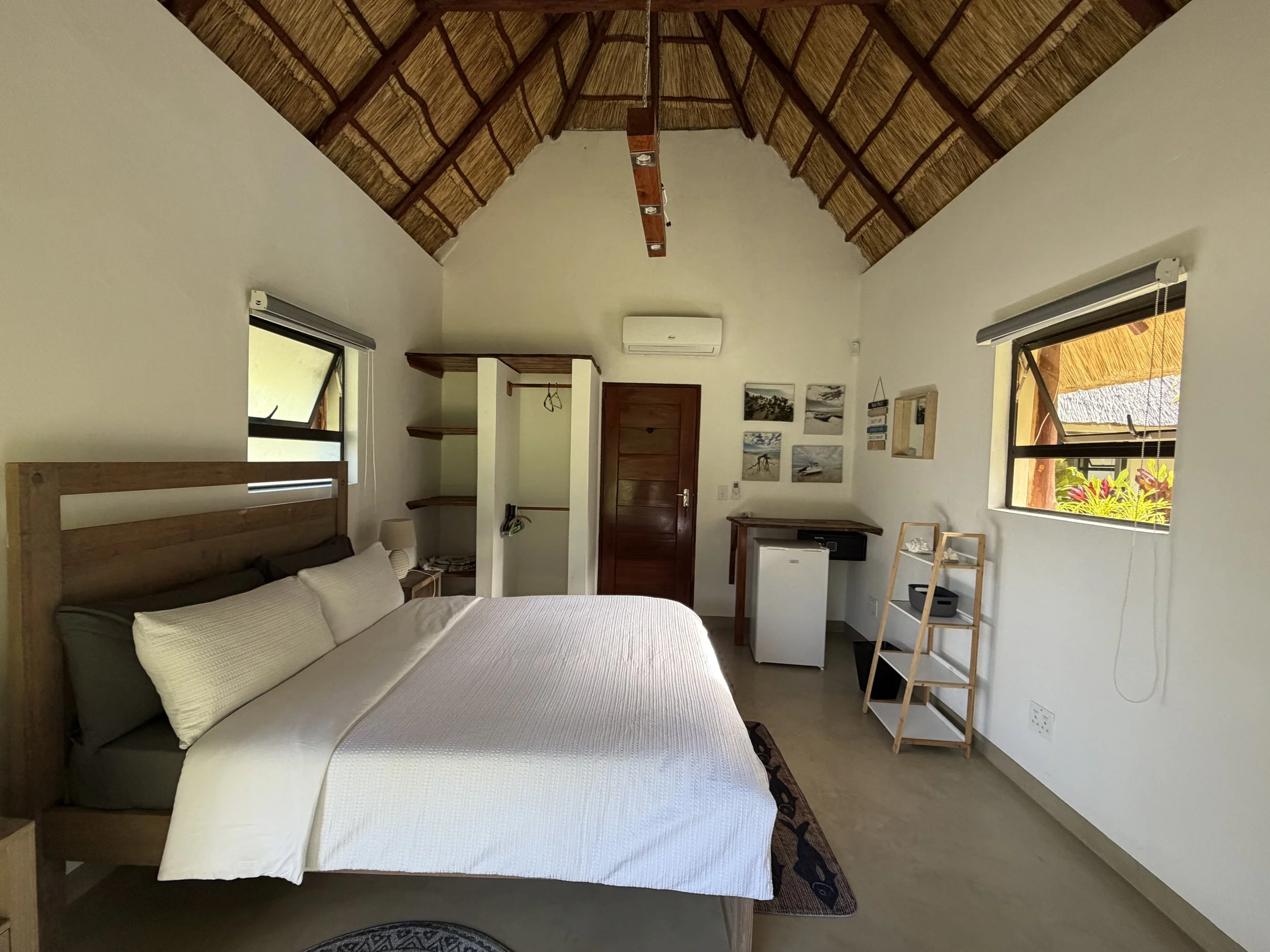A bedroom with a sloped thatched ceiling, large window on the right, and a bed with white bedding and pillows in the lower left corner. There are wall-mounted pictures on the wall, a small fridge, a ladder-style shelf, and an air conditioner.