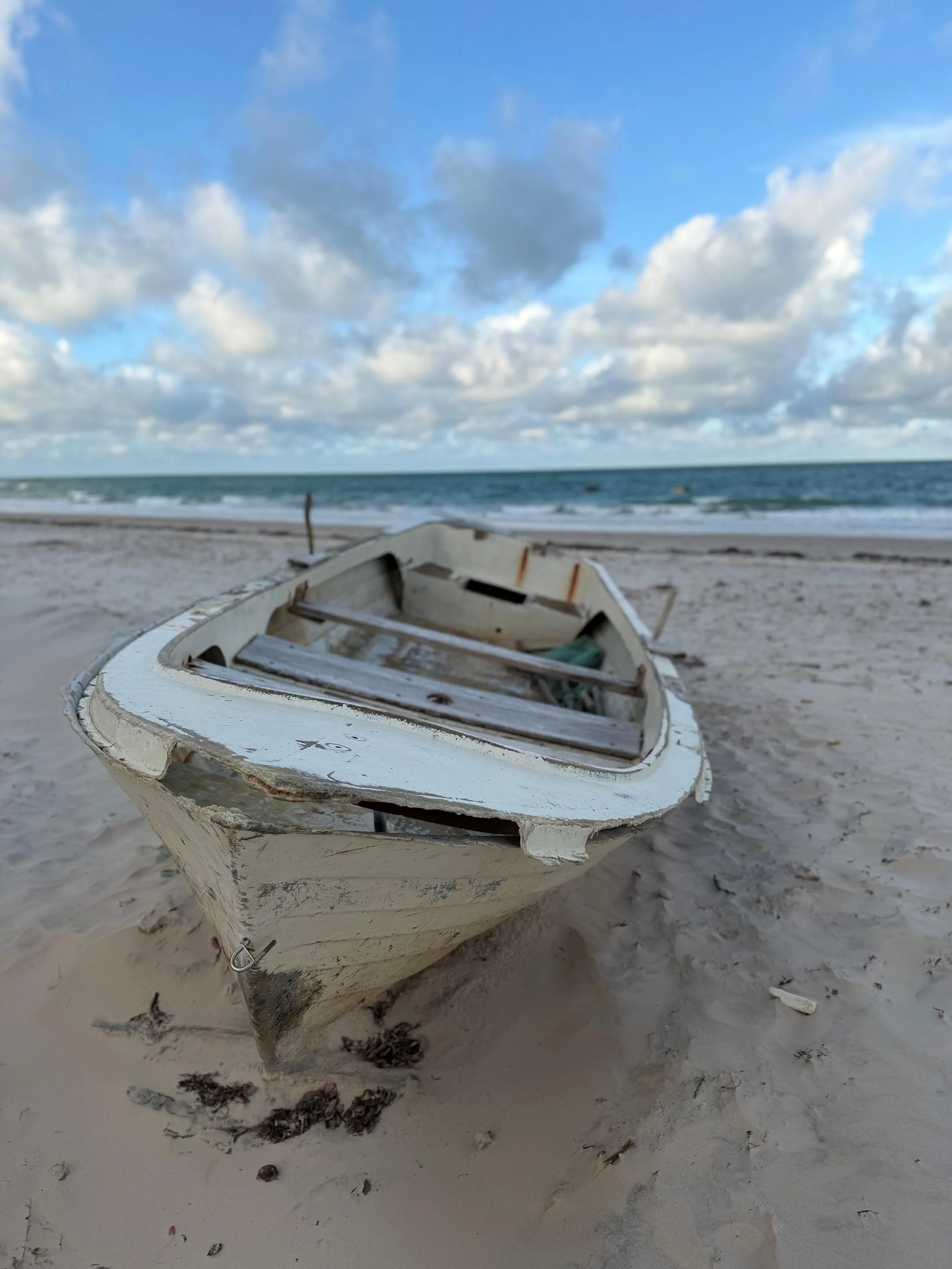 An old, weathered white rowboat on a sandy beach with ocean waves and a blue sky with clouds in the background.
