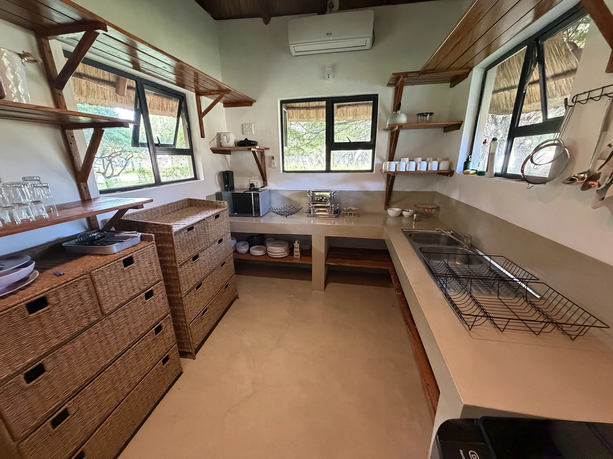 Kitchen with wooden shelves, a microwave, dishes, and windows showing greenery outside.