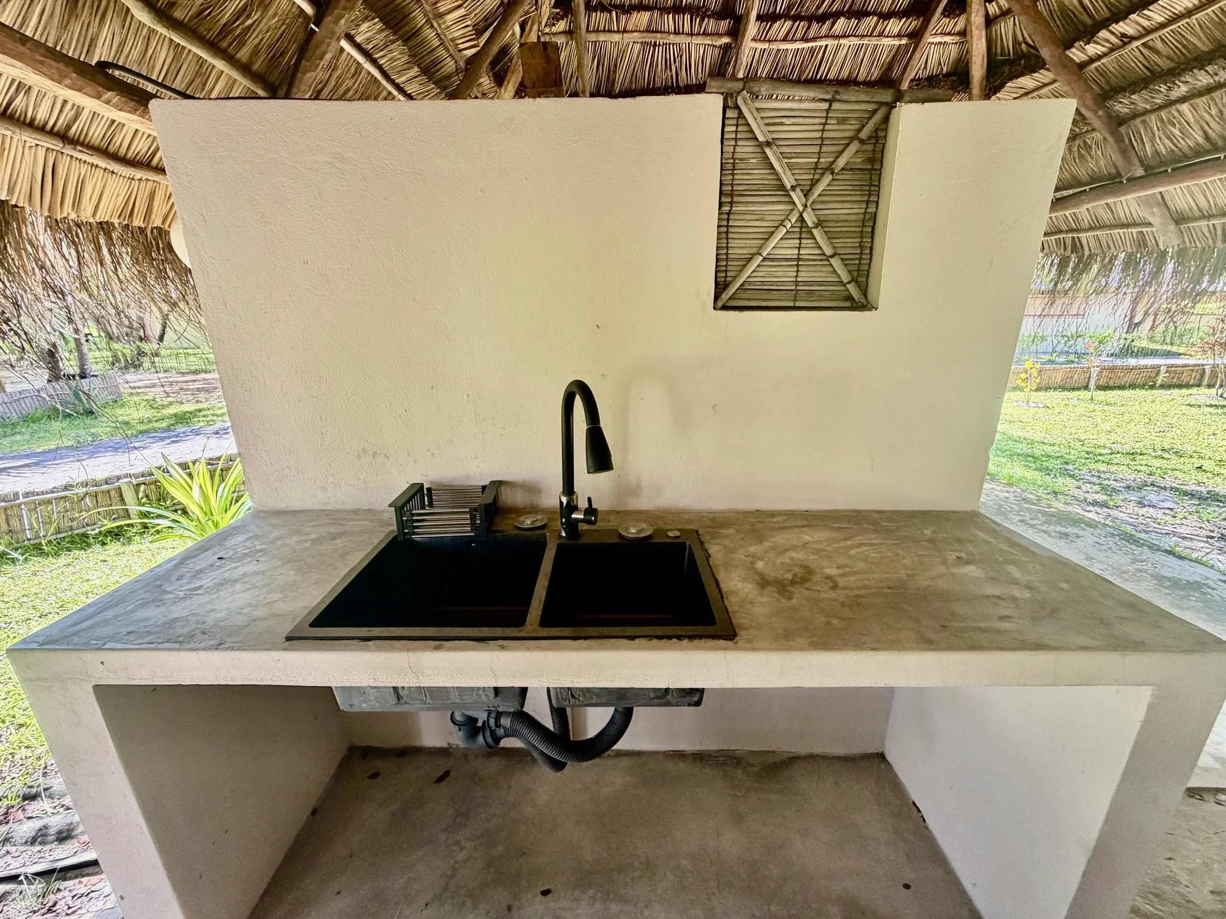 The image shows a kitchen sink with a black faucet, set on a stone countertop, beneath a white roof with a straw thatched ceiling. There are plants visible outside through open spaces in the roof structure.