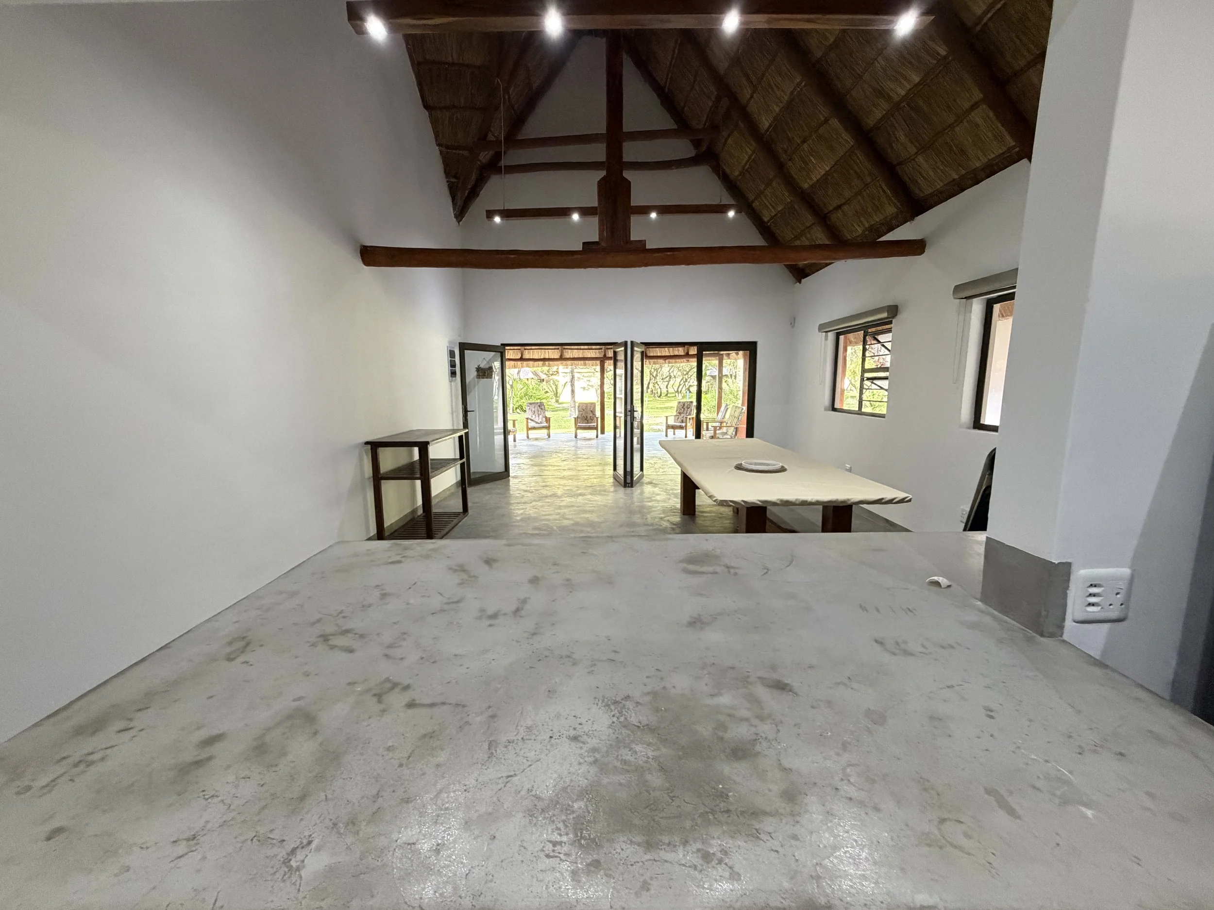 Interior of a room with a high, thatched, vaulted ceiling with exposed wooden beams. The room opens to an outdoor patio with chairs and a garden view.