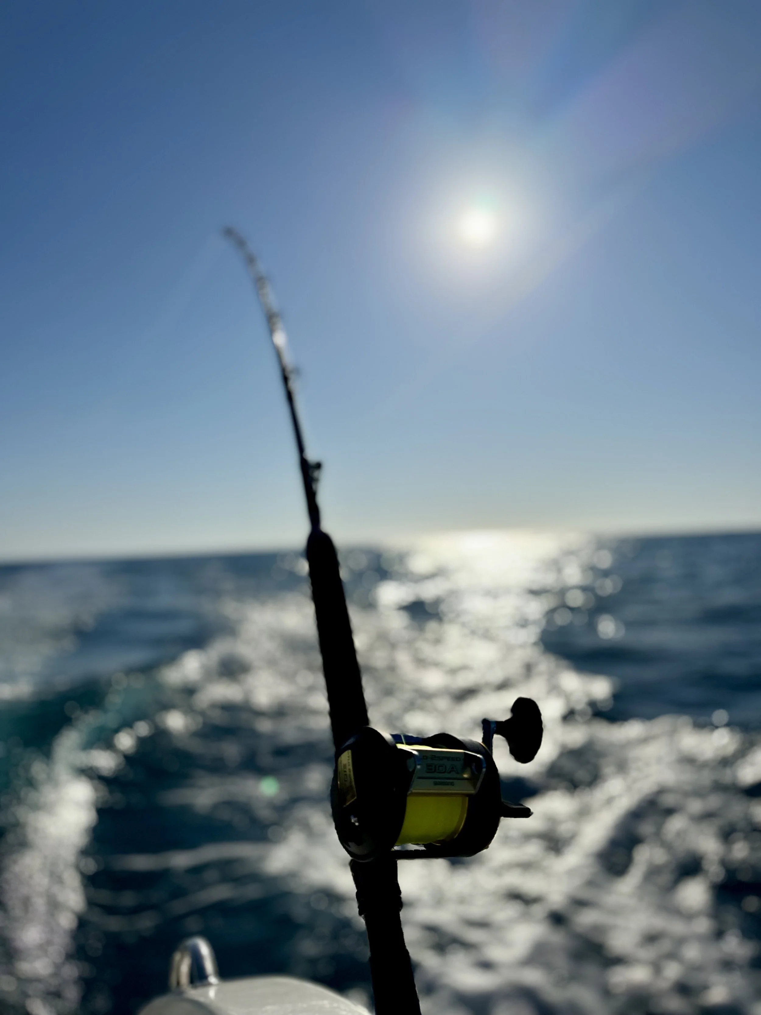 A close-up view on a boat with a fishing rod extending into the water, sunlight reflecting on the ocean surface, and a clear blue sky.
