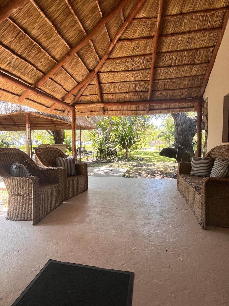 Outdoor covered patio with wicker furniture and striped cushions, surrounded by trees and lush greenery.