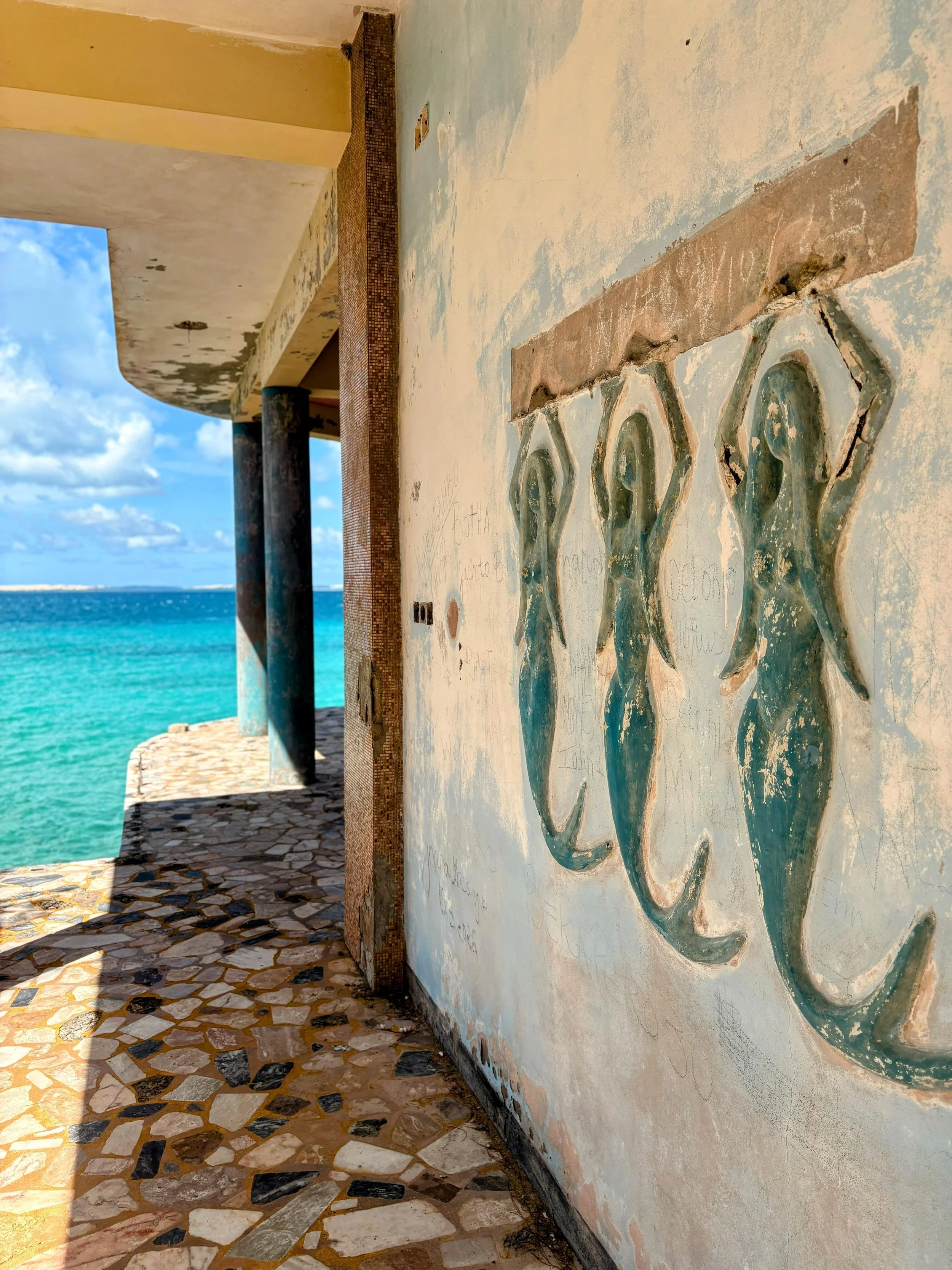 A weathered wall with painted artwork of dolphins, located on a balcony overlooking the ocean, with a mosaic-tiled floor and support columns, under a partly cloudy sky.