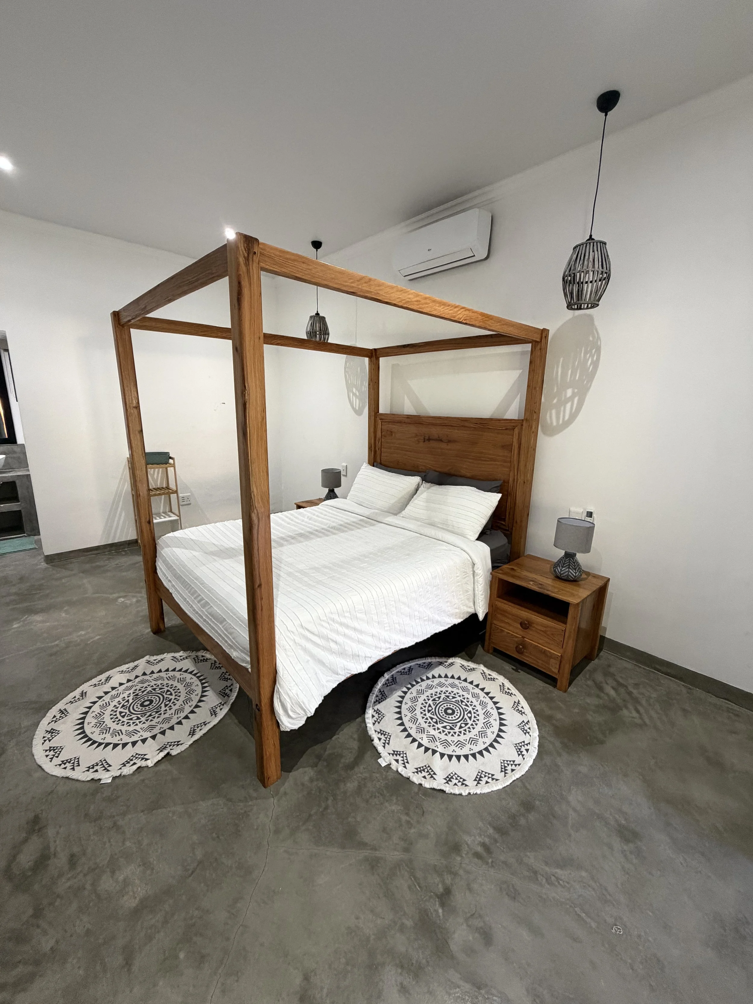 A bedroom with a wooden canopy bed, white bedding, two small round black and white patterned rugs on a concrete floor, a nightstand with a gray lamp, and two black pendant lights hanging from the ceiling. There is an air conditioning unit on the wall.