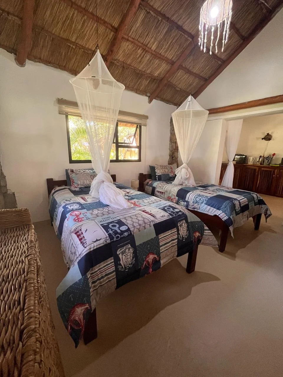 Two twin beds with patchwork quilt covers and white mosquito nets hanging over each bed in a rustic, tropical-styled bedroom with a thatched ceiling and a window with natural light.