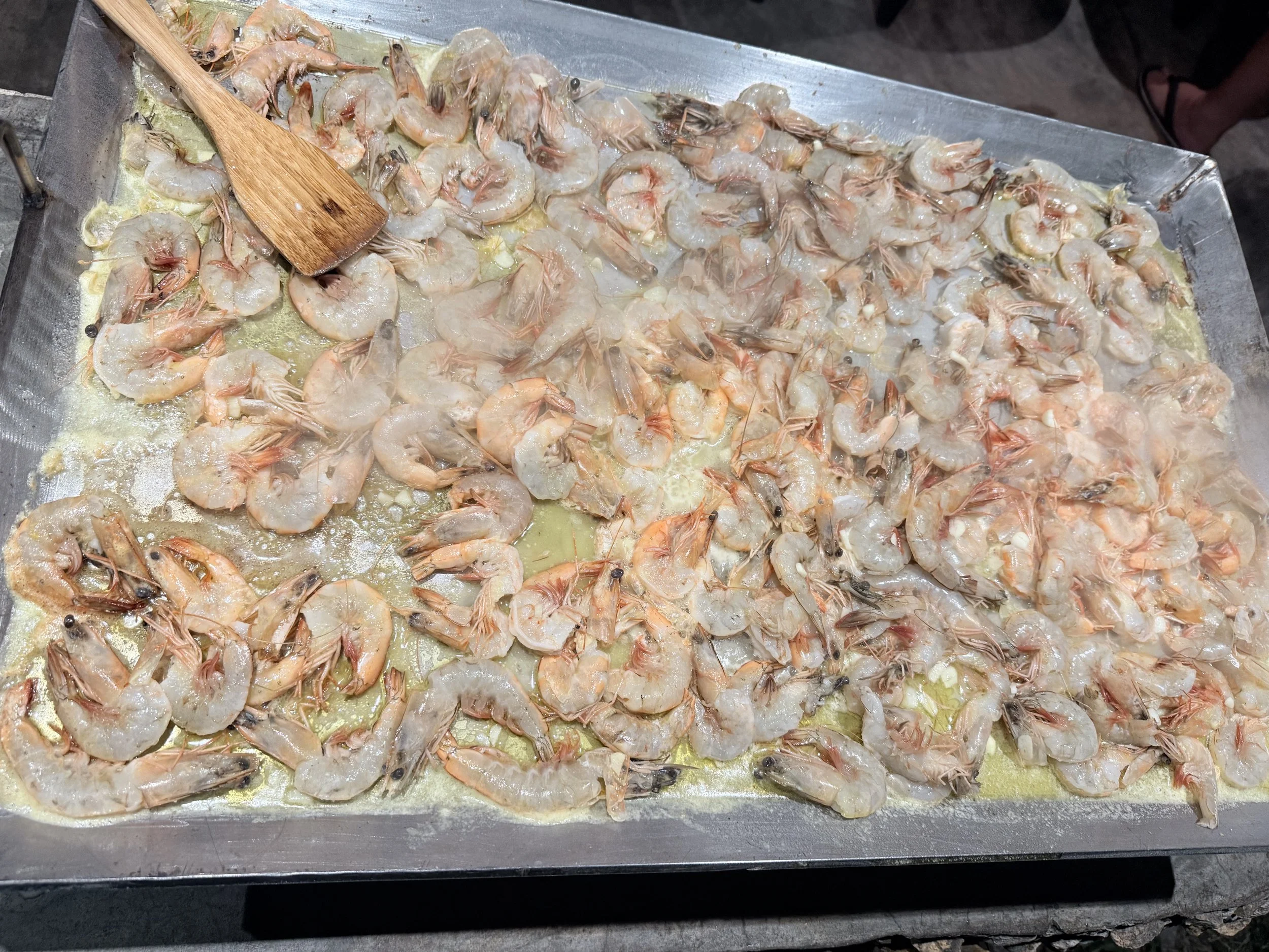 Raw shrimp cooking on a metal tray with a wooden spatula, with visible butter or oil in the tray.