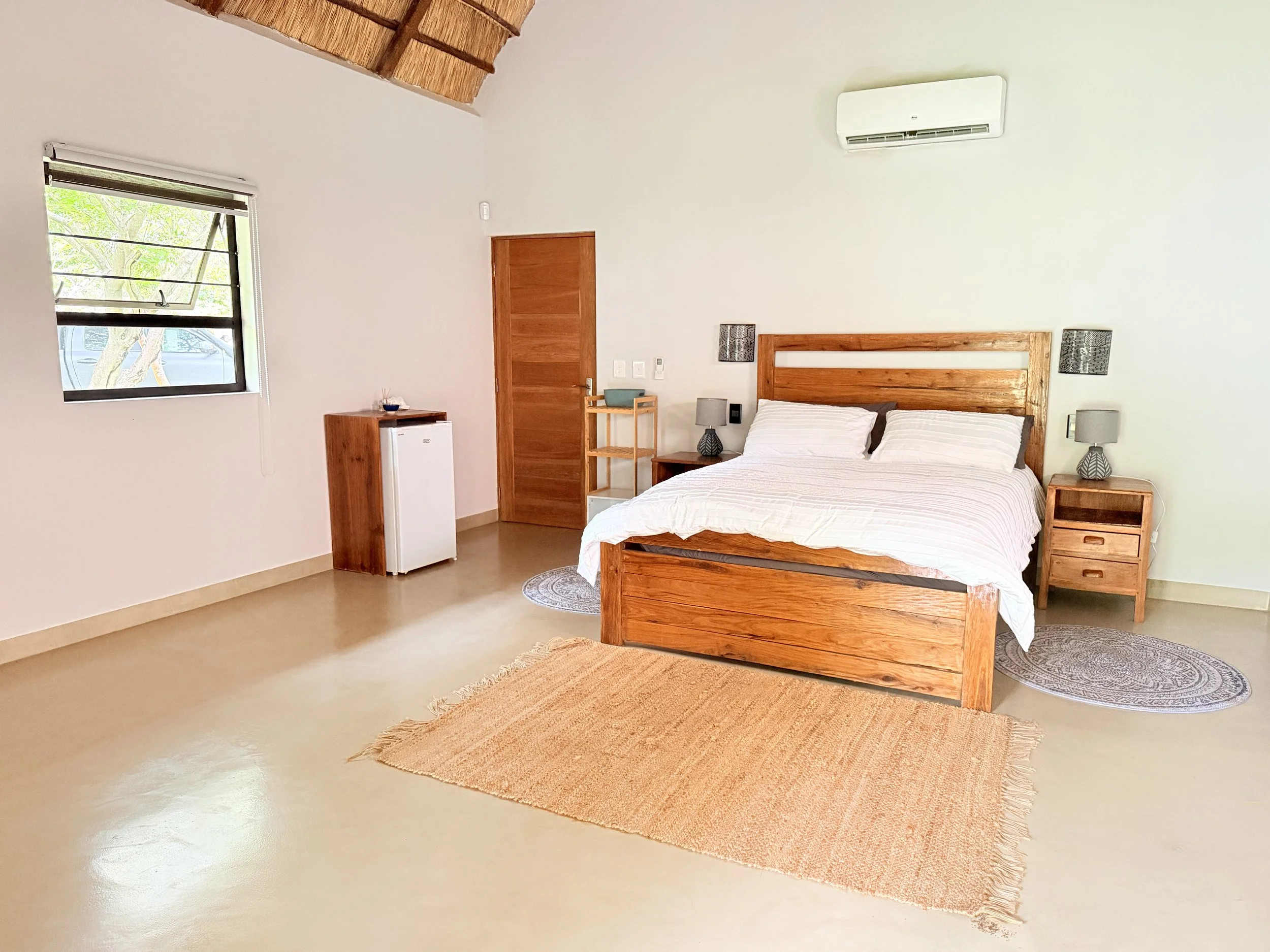 A bedroom with a wooden bed, two nightstands with matching lamps, a small rug, a window, a wooden door, and a white mini refrigerator. The room has a high vaulted ceiling with wooden beams and an air conditioning unit on the wall.