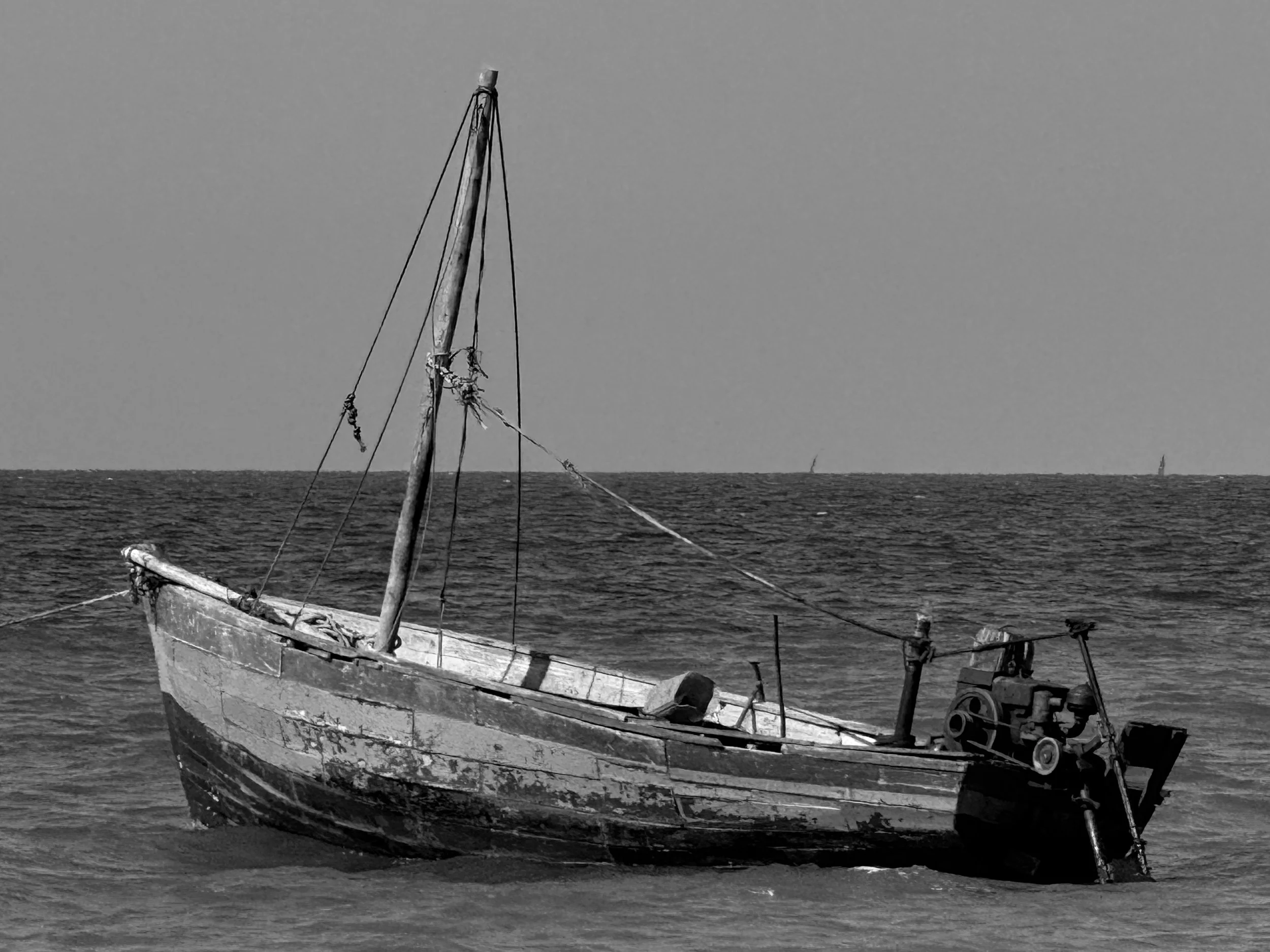An old, weathered boat with peeling paint sitting on the water, with a tugboat in the background and a cloudy sky.