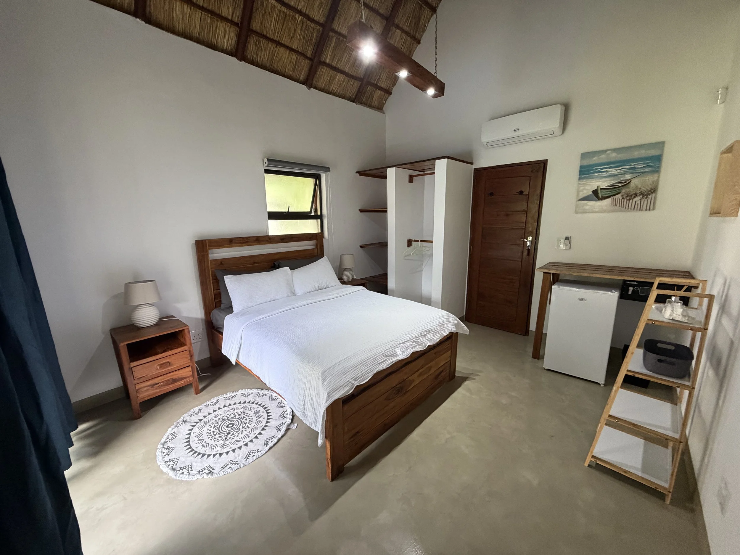 A room with a wooden bed frame and white bedding, two nightstands with lamps, a circular patterned rug, a small window, a white open wardrobe, wooden shelves, an air conditioning unit, a door, a wooden table with a white mini fridge underneath, and a wooden drying rack.