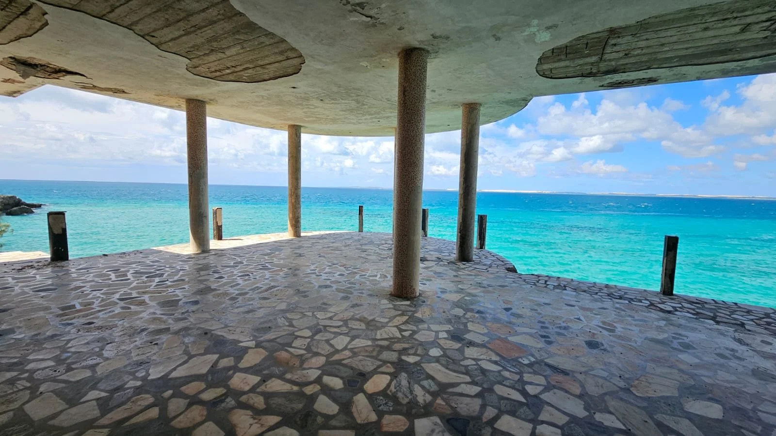 Underwater bar or lounge with a stone floor, concrete ceiling, and wooden support columns overlooking turquoise ocean and partly cloudy sky.