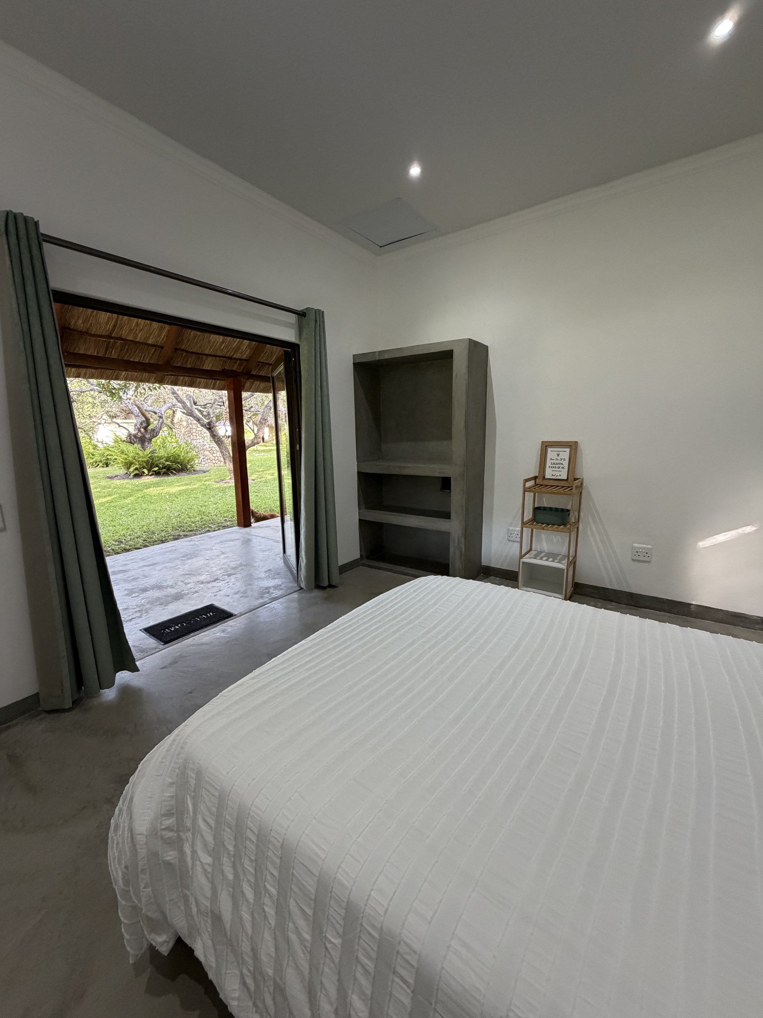 A bedroom with a white bedspread, an open sliding glass door leading to an outdoor patio with a thatched roof, and a view of trees and greenery outside.