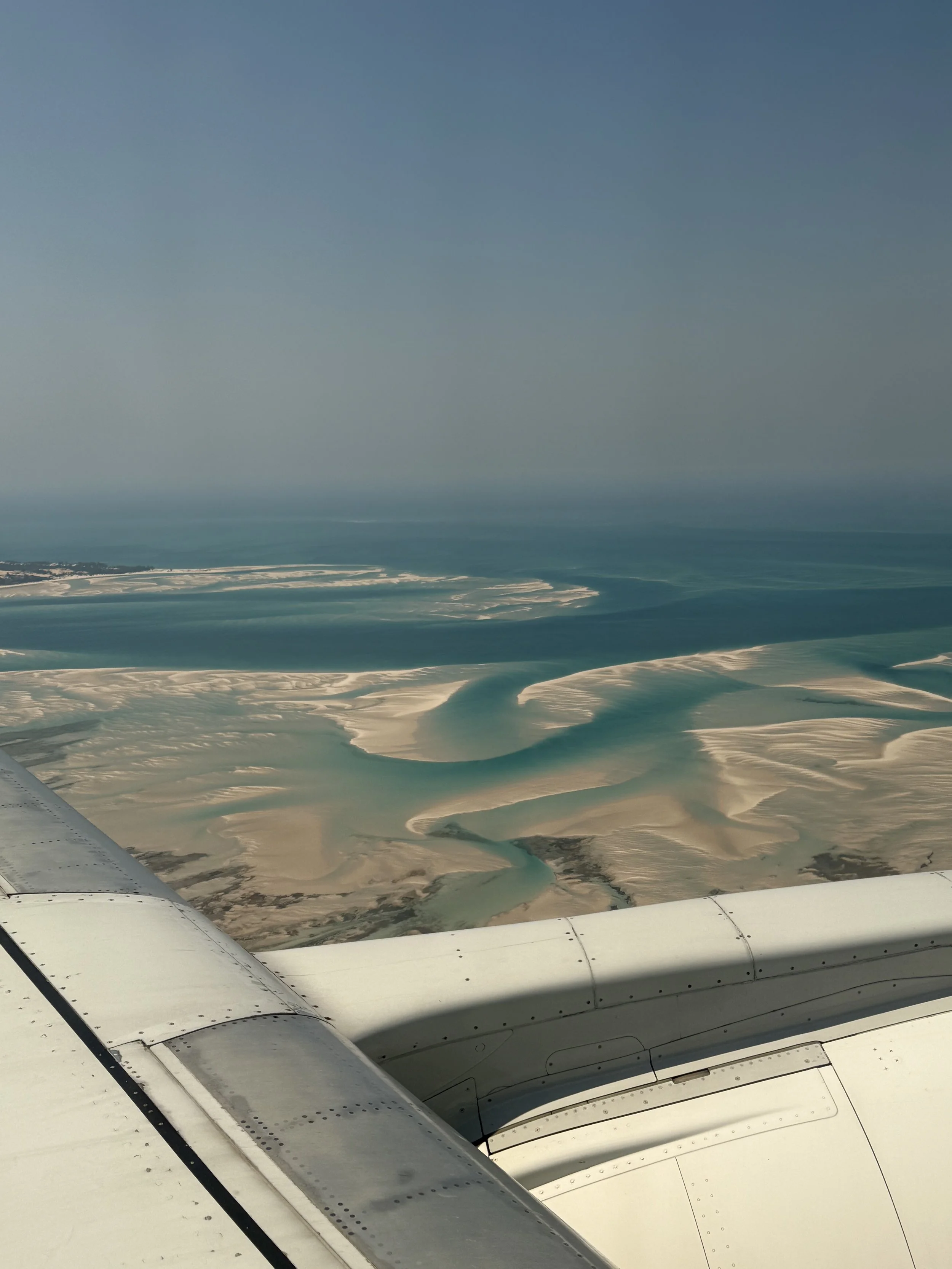 View from an airplane window showing the aircraft's wing and a landscape below with winding rivers and sand or salt flats, under a clear blue sky.