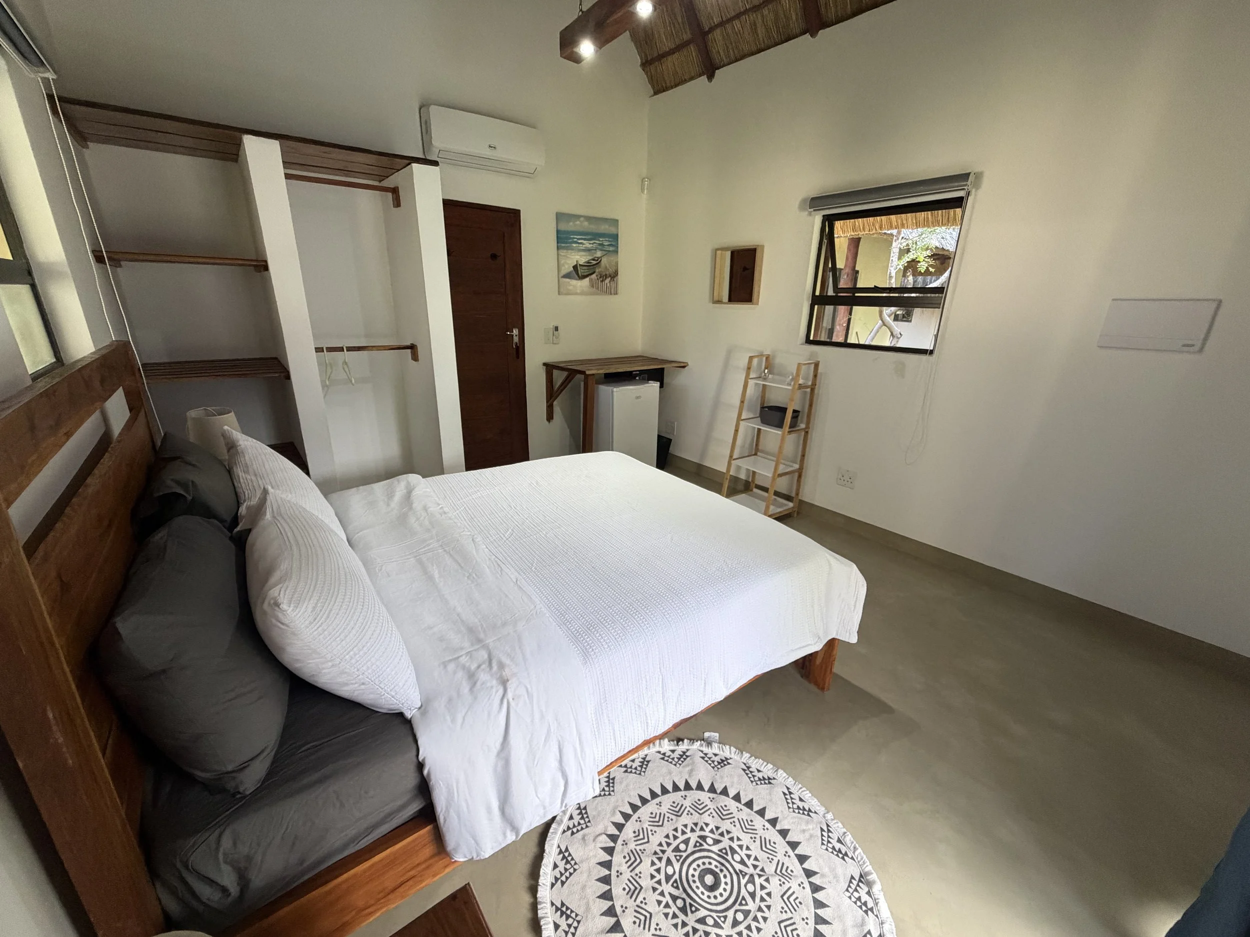 A bedroom with a large bed covered with white bedding and pillows, a circular black and white patterned rug, a small wooden bookshelf, a window with partially open blinds showing trees outside, a small table, and wall art depicting a boat near the beach.