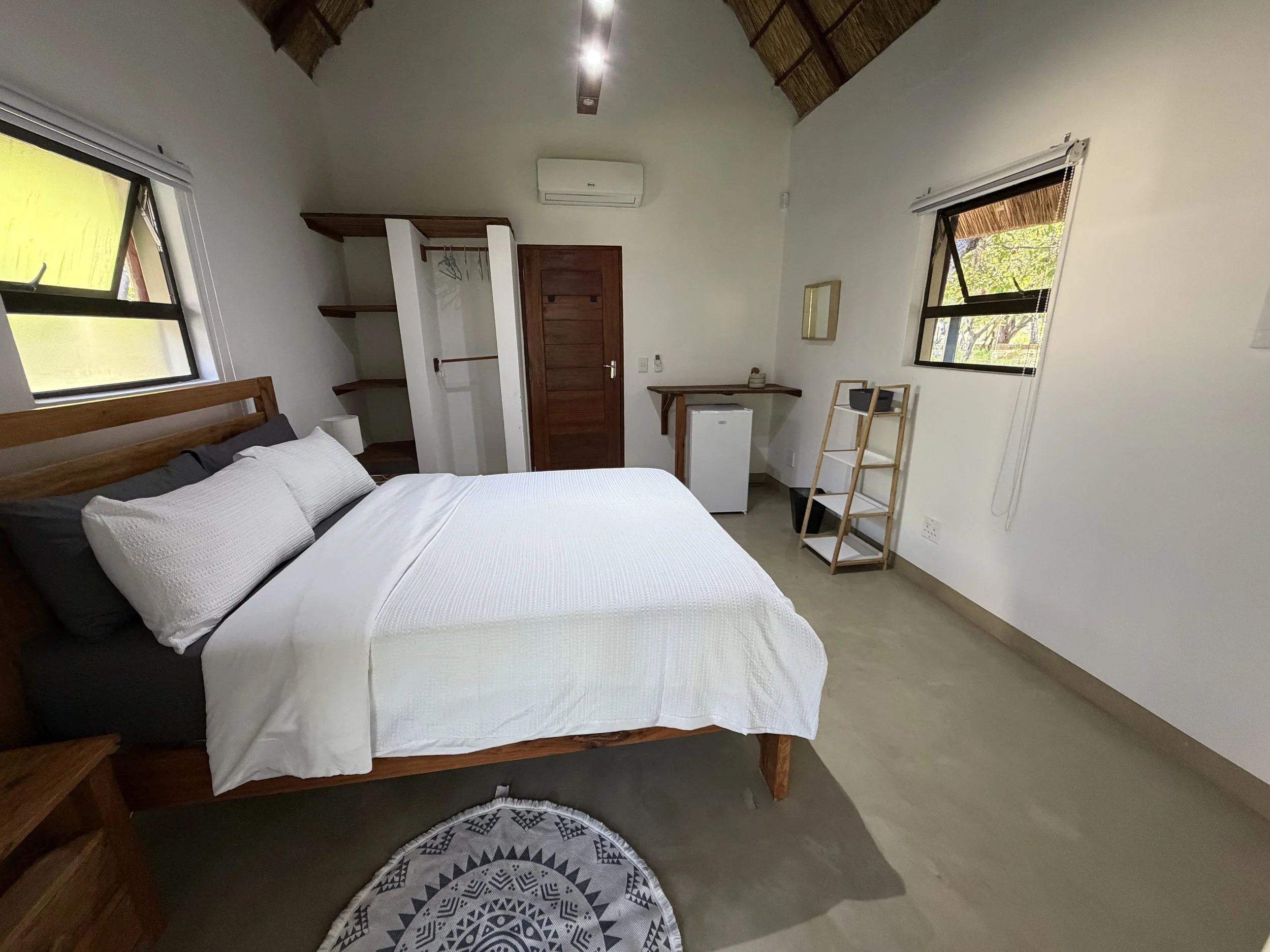Interior of a minimalist bedroom with a bed featuring white bedding, two windows with black frames and bamboo shades, an air conditioning unit, a small wooden shelf with a plant, and a patterned round rug.