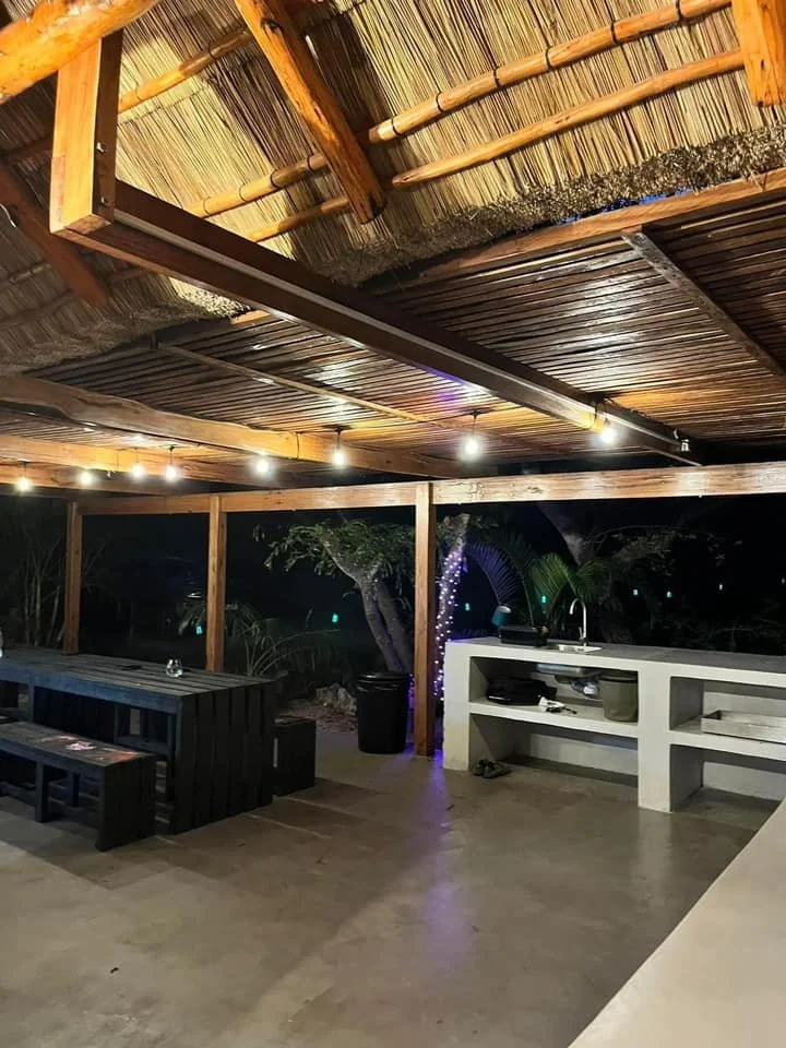 An outdoor patio area with a wooden roof supported by wooden beams, string lights hanging from the ceiling, a black bench, a white counter with a sink, and some greenery in the background.