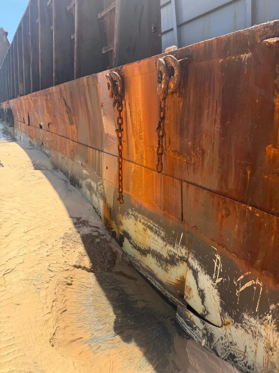 Rusty metal ship hull on sandy ground with chains hanging from the side.