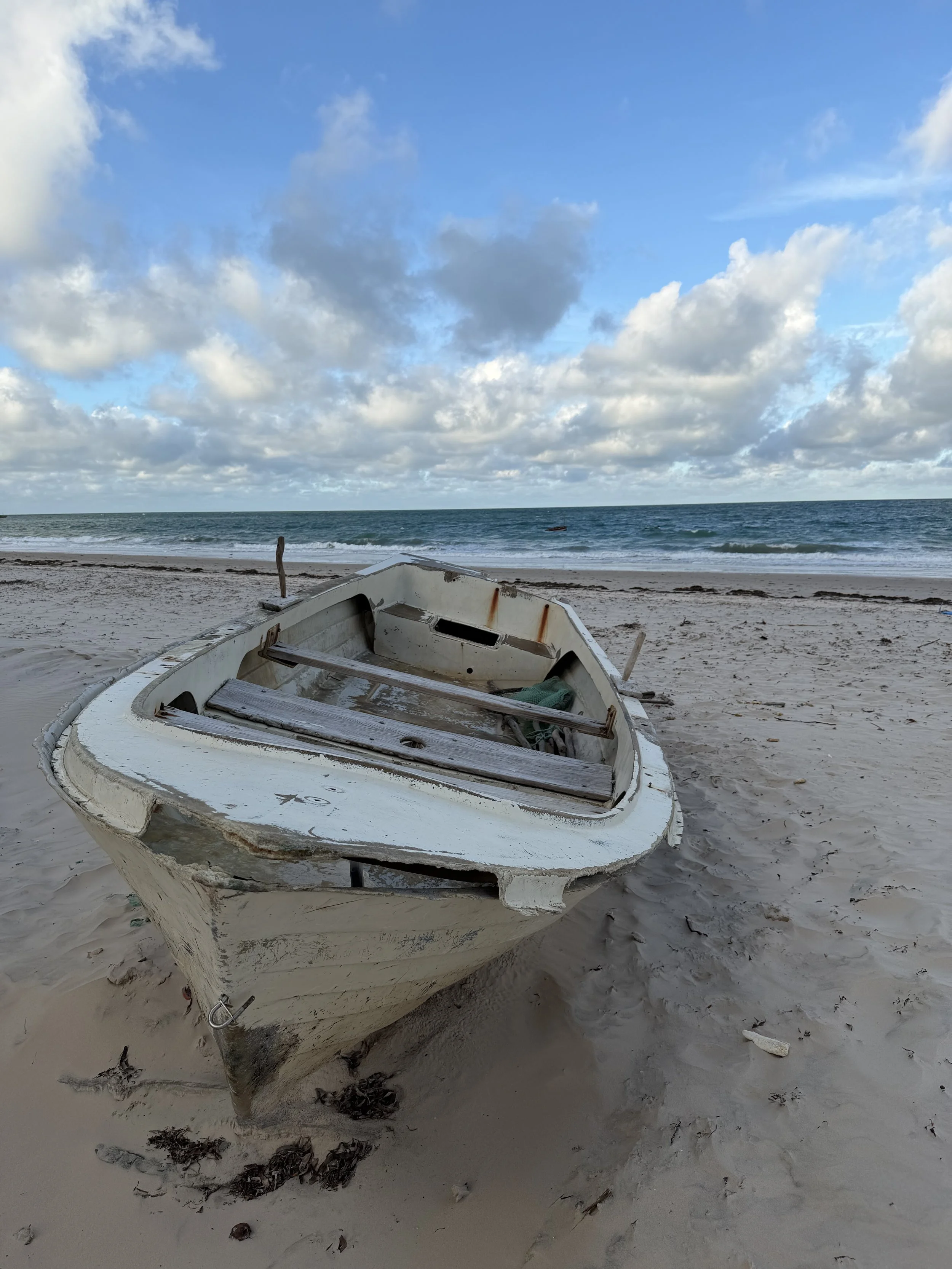 An abandoned, broken boat on a sandy beach with ocean and cloudy sky in the background.