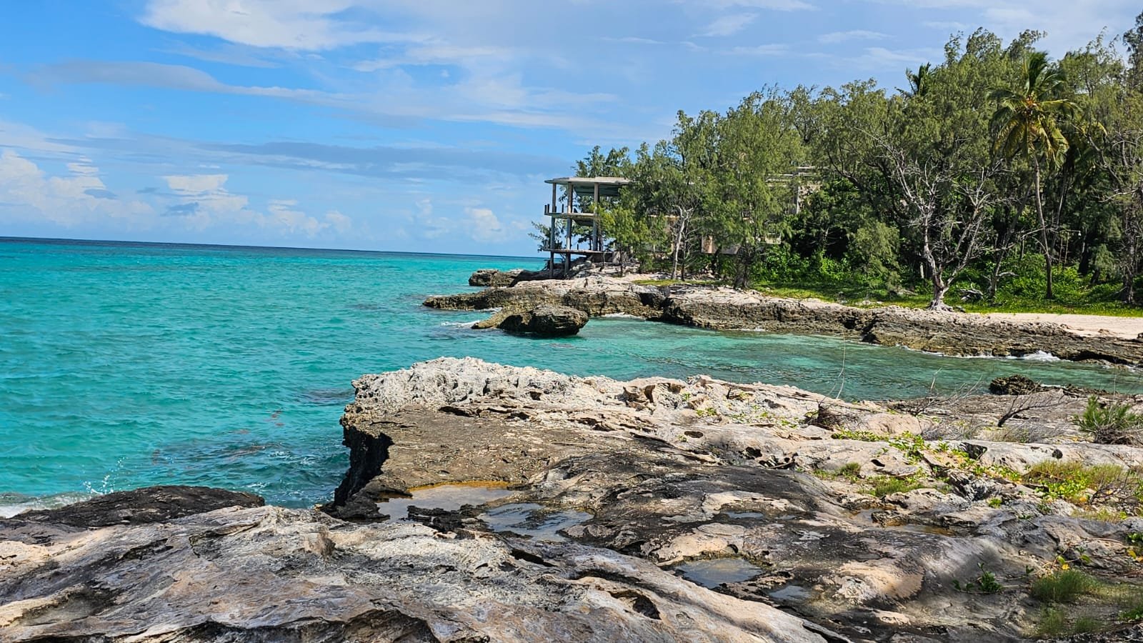 Tropical beach with turquoise water, rocky shoreline, green trees, and a modern house on stilts overlooking the ocean.