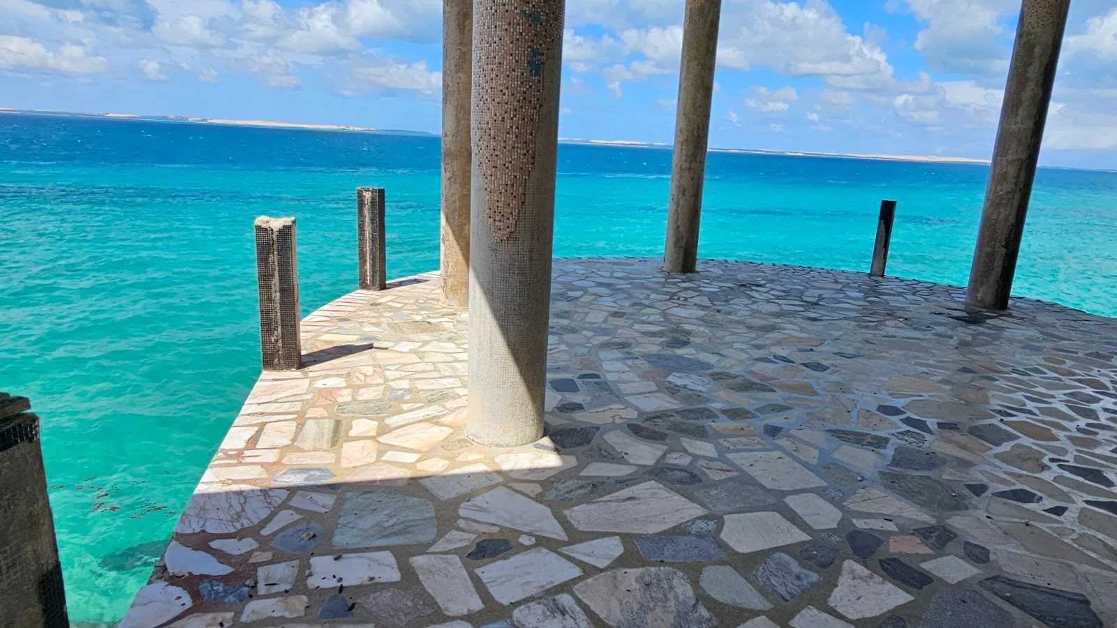 A stone-tiled platform with large cylindrical pillars extends over turquoise ocean water under a partly cloudy sky.