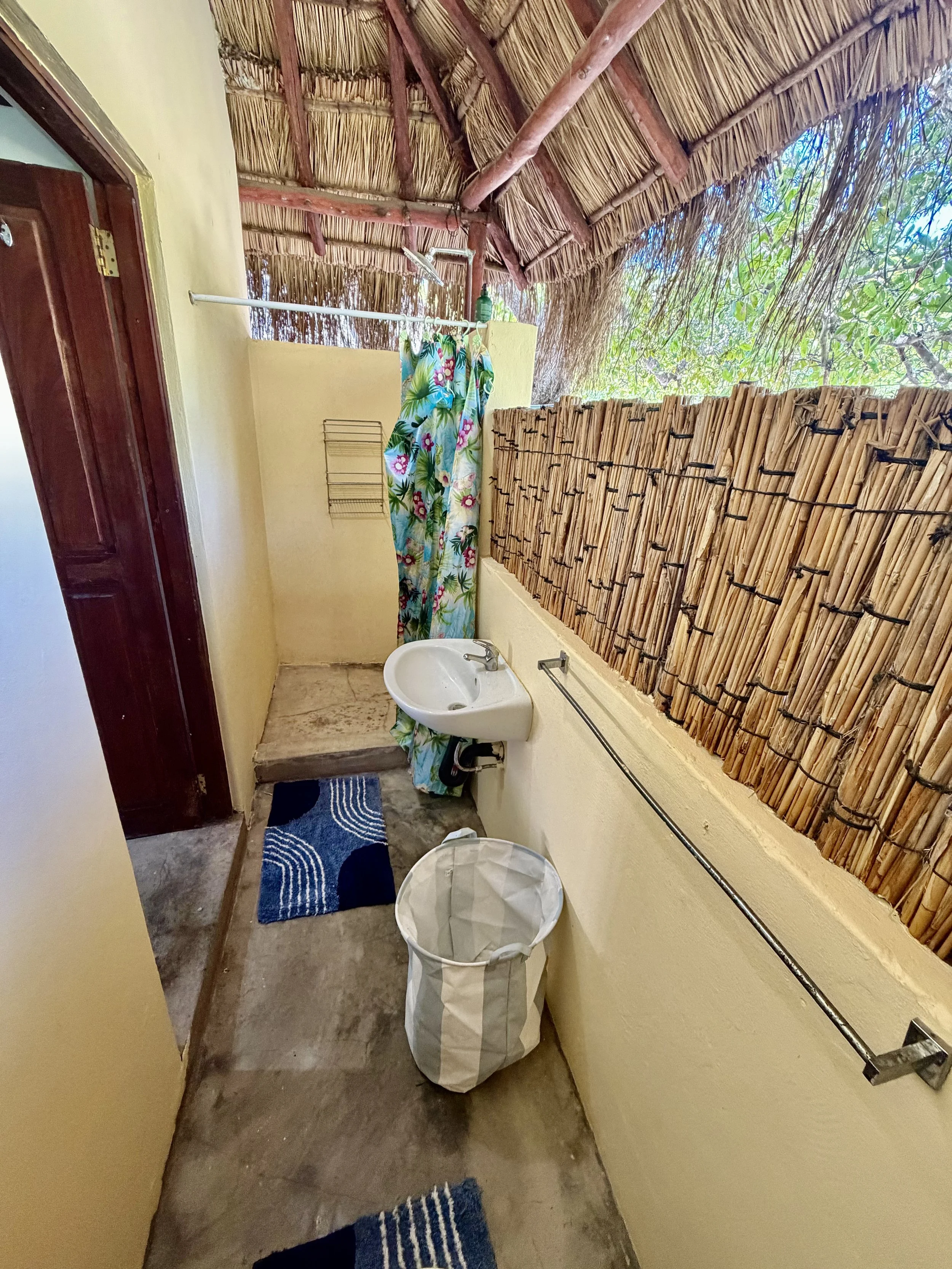 Bathroom with yellow walls, bamboo privacy screen, small white sink, blue and black striped rugs, laundry basket, and hanging floral shower curtain under a thatched roof ceiling.