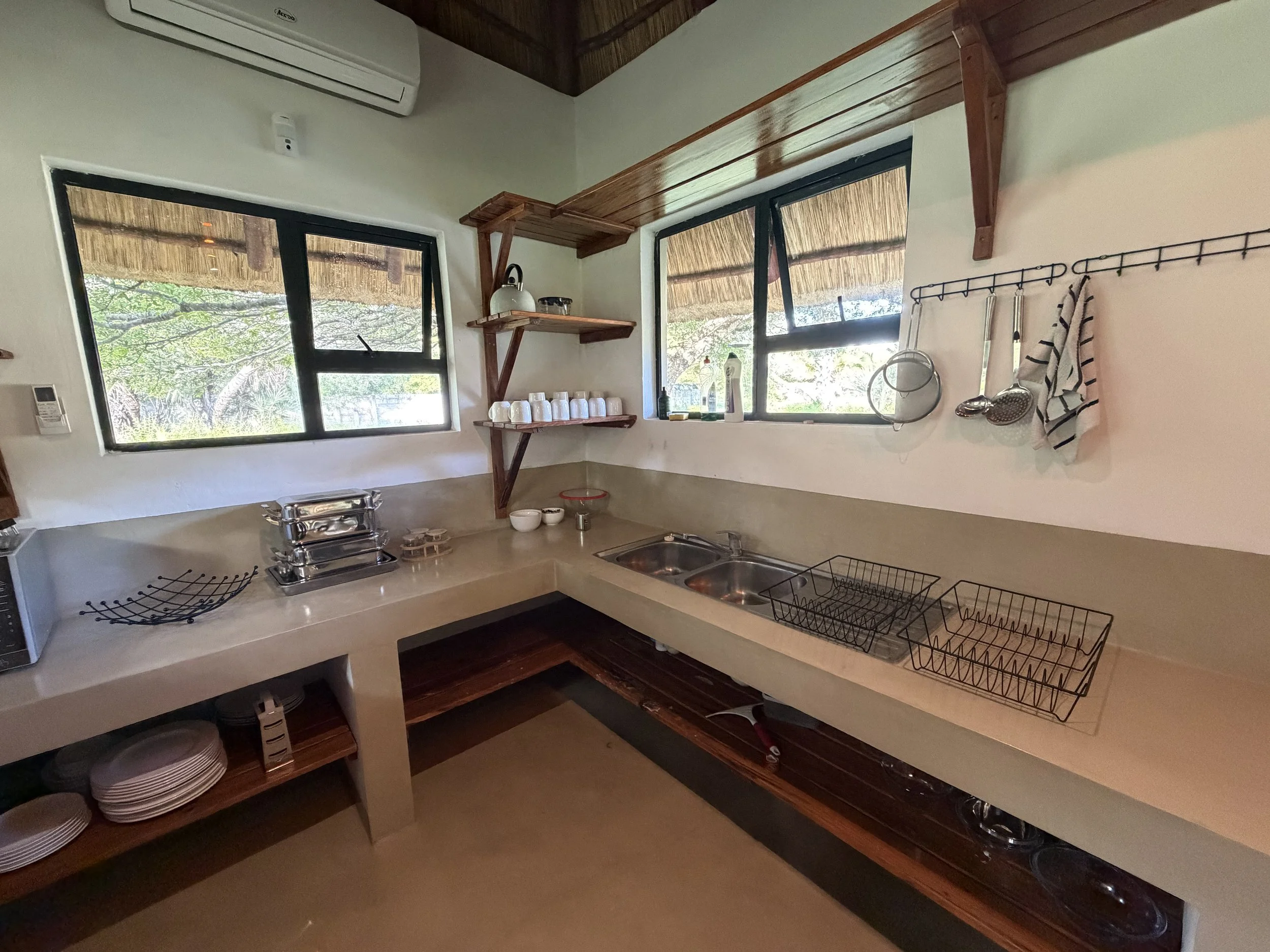 Kitchen with two windows, wooden shelves, and white walls. Kitchen counter with dishes, utensils, and a dish rack.