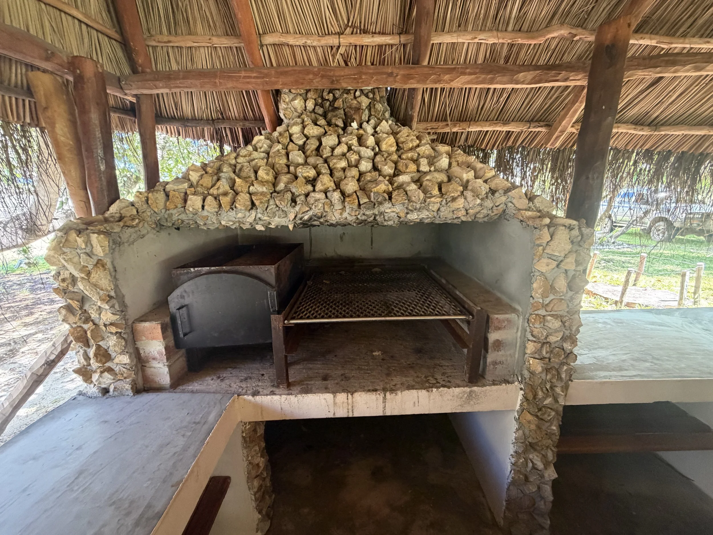 A stone and brick outdoor fireplace with a thatched roof and wooden beams, situated in a tropical setting.