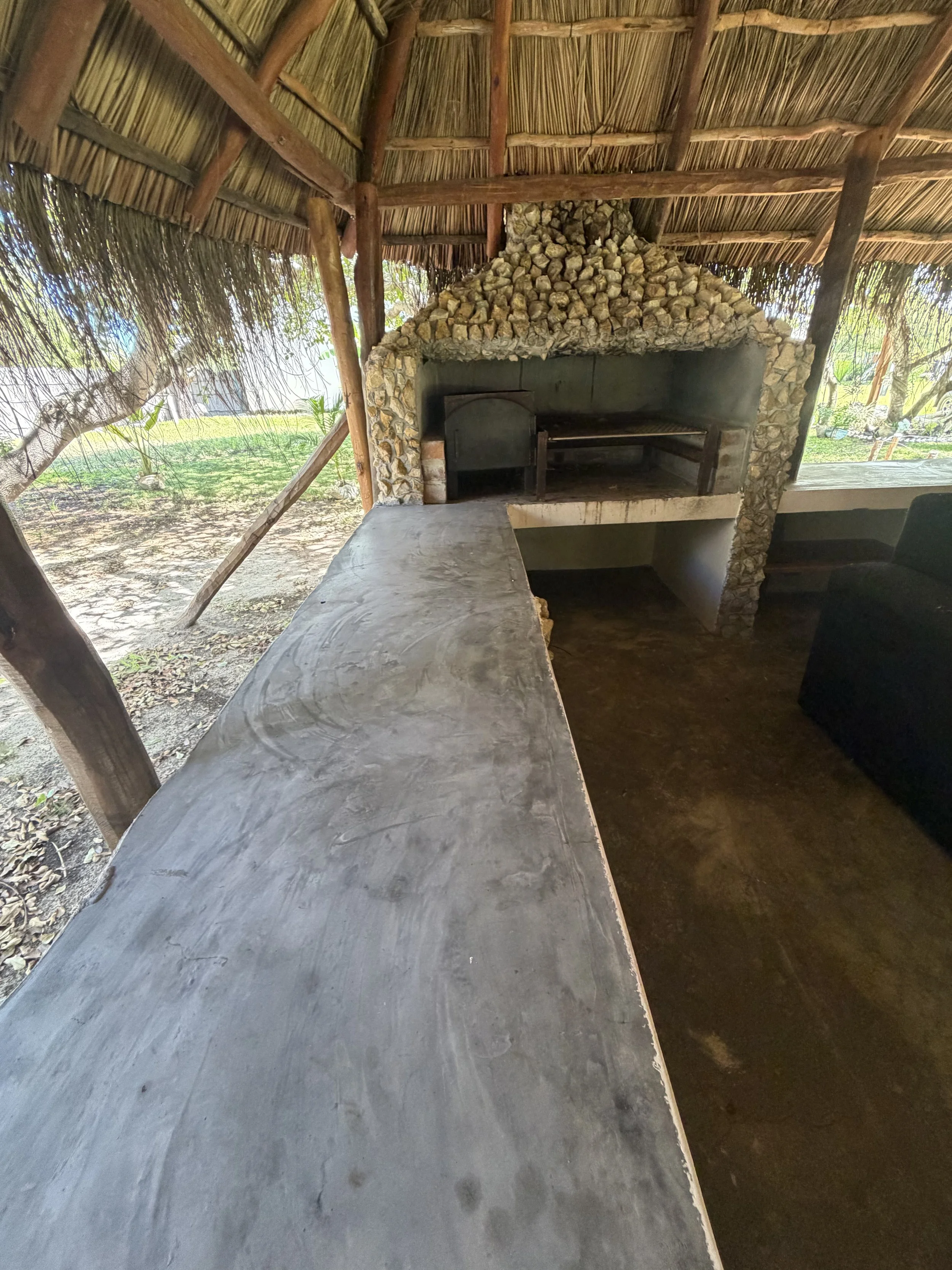 Barbecue or outdoor kitchen area with a stone oven and a concrete countertop, surrounded by a thatched roof structure and natural outdoor scenery.