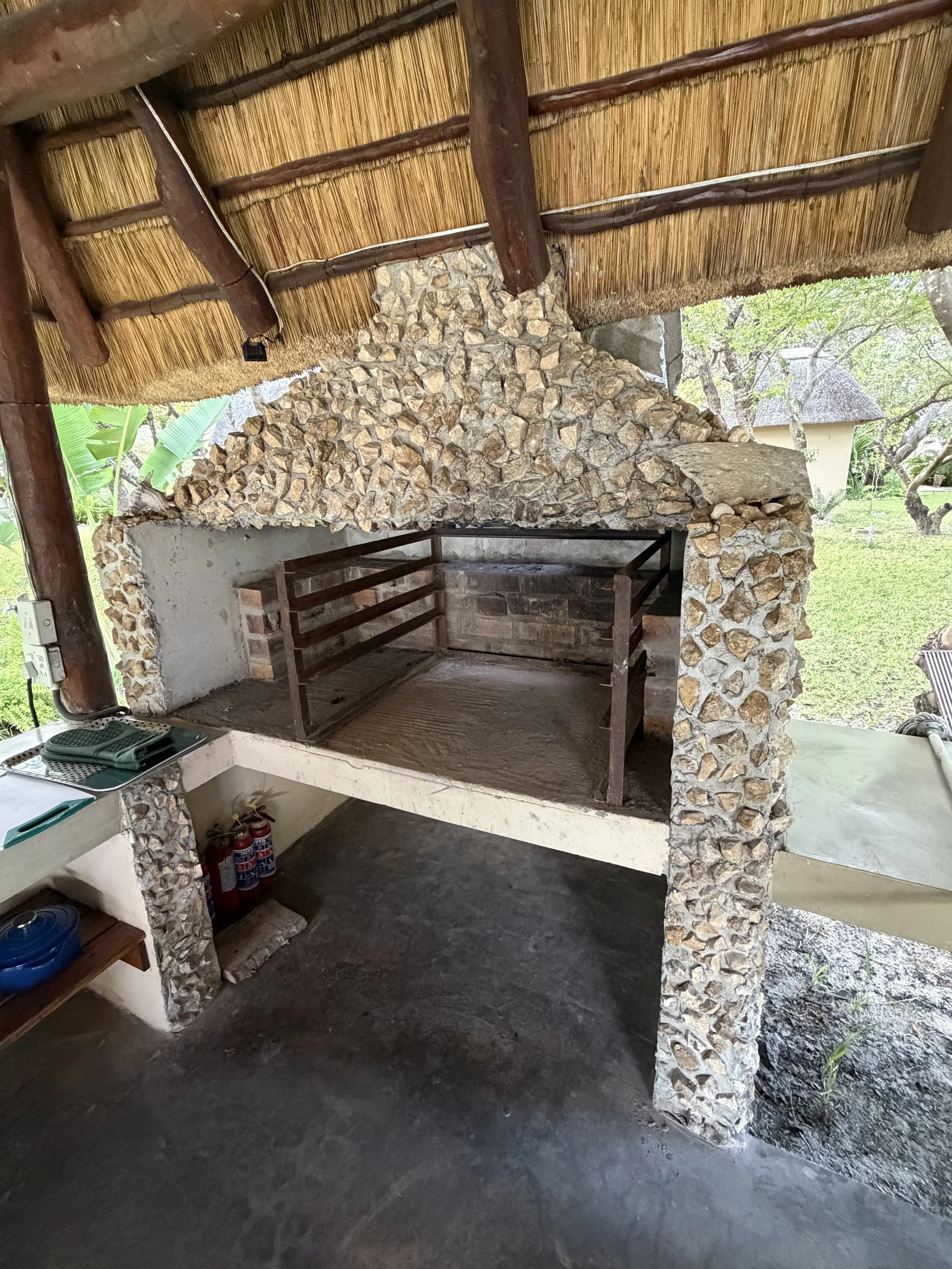 A rustic outdoor fireplace with a stone structure and a wooden frame, located in a garden setting with trees and grass visible in the background.