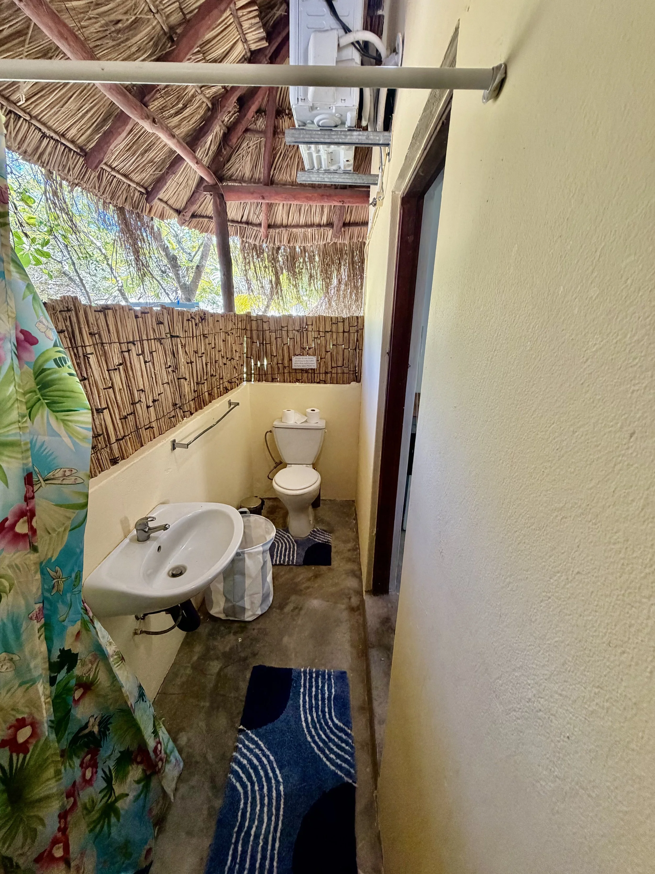 Small bathroom with a yellow wall, a toilet, a sink, and a blue patterned rug. There is a tropical shower curtain with floral and leaf designs. The bathroom has a thatched roof ceiling and a bamboo privacy wall outside the window.