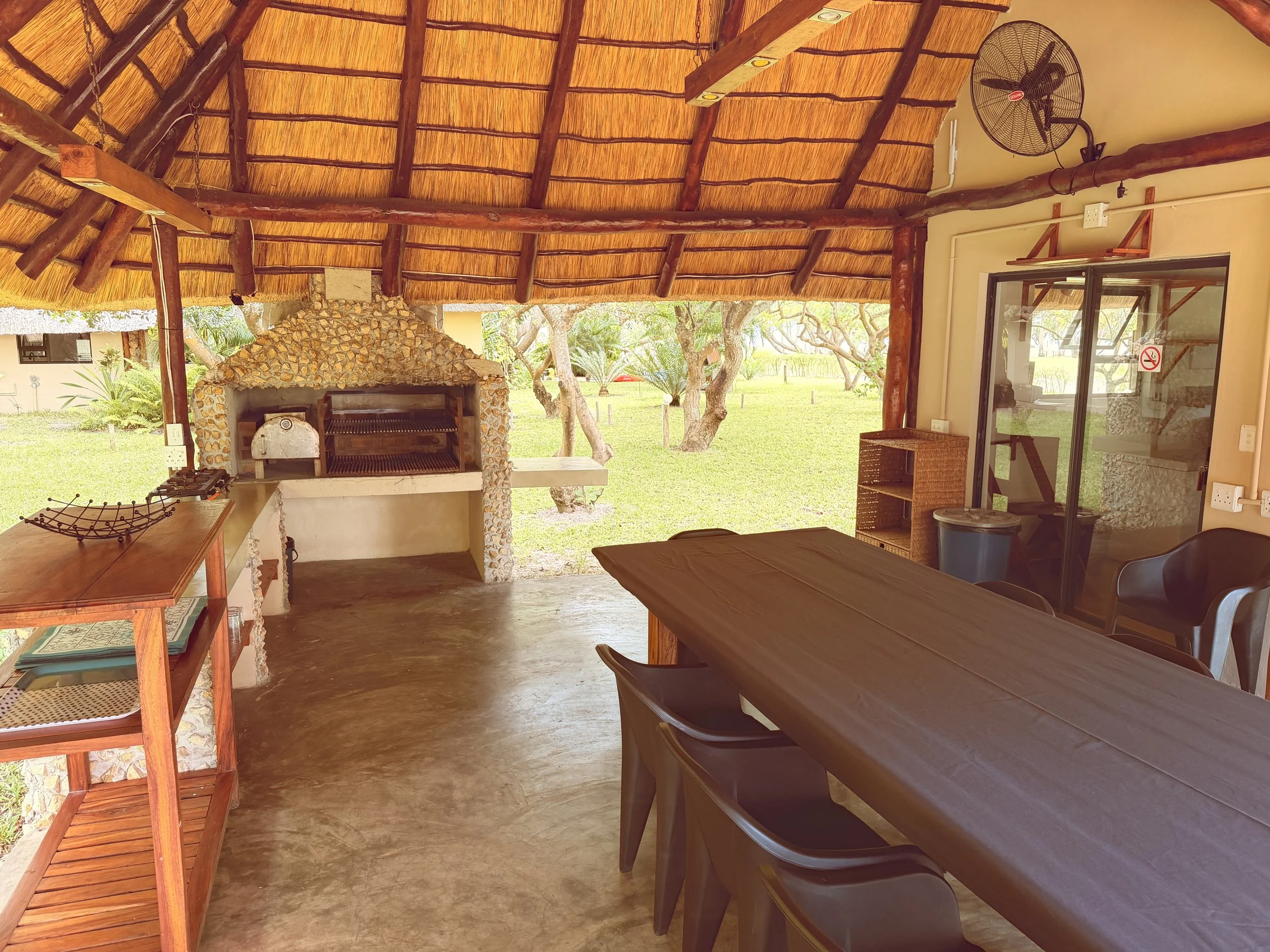 View of a covered outdoor patio area with a thatched roof, a stone barbecue grill, benches, tables, and outdoor surroundings with trees and grass.