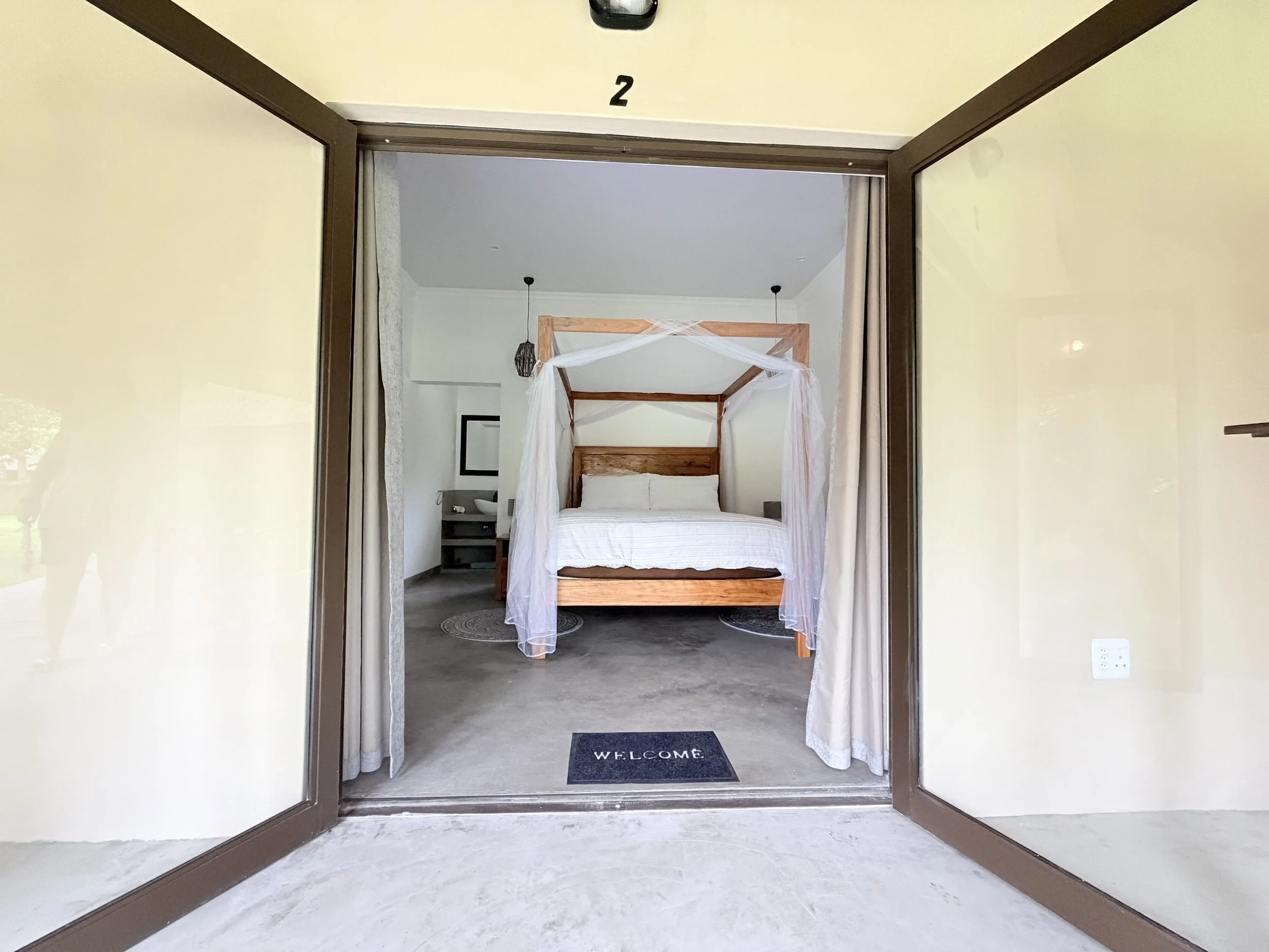 Open door leading into a bedroom with a four-poster bed and white bedding, decorated with sheer curtains. A black welcome mat is on the floor outside the door.