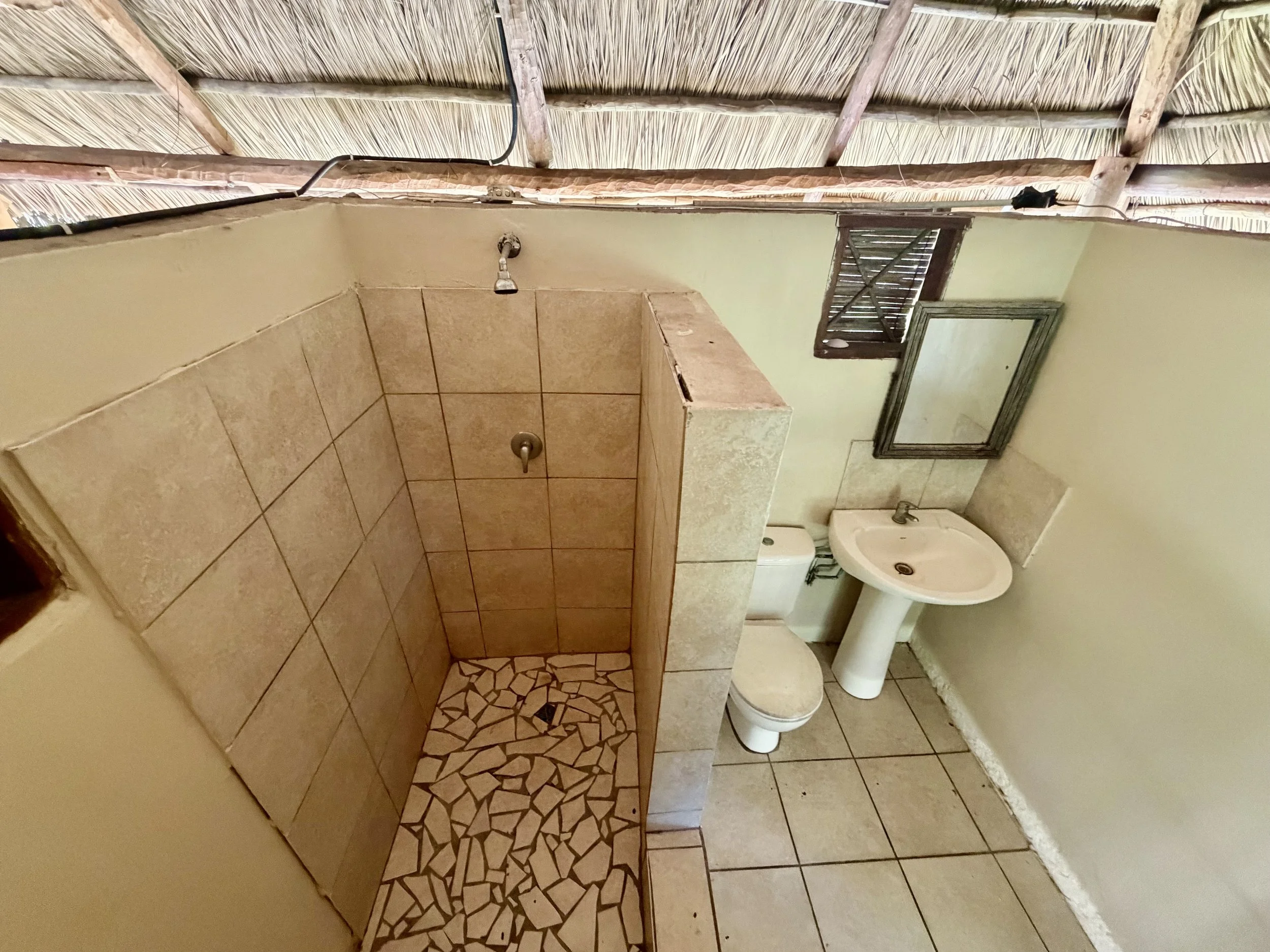 Inside a bathroom featuring tiled walls and floors, a small window with wooden slats, a pedestal sink, a toilet, and a terrazzo-style tiled walk-in shower area with beige and brown tiles. The ceiling appears to be low, with exposed wooden beams and thatched roofing.