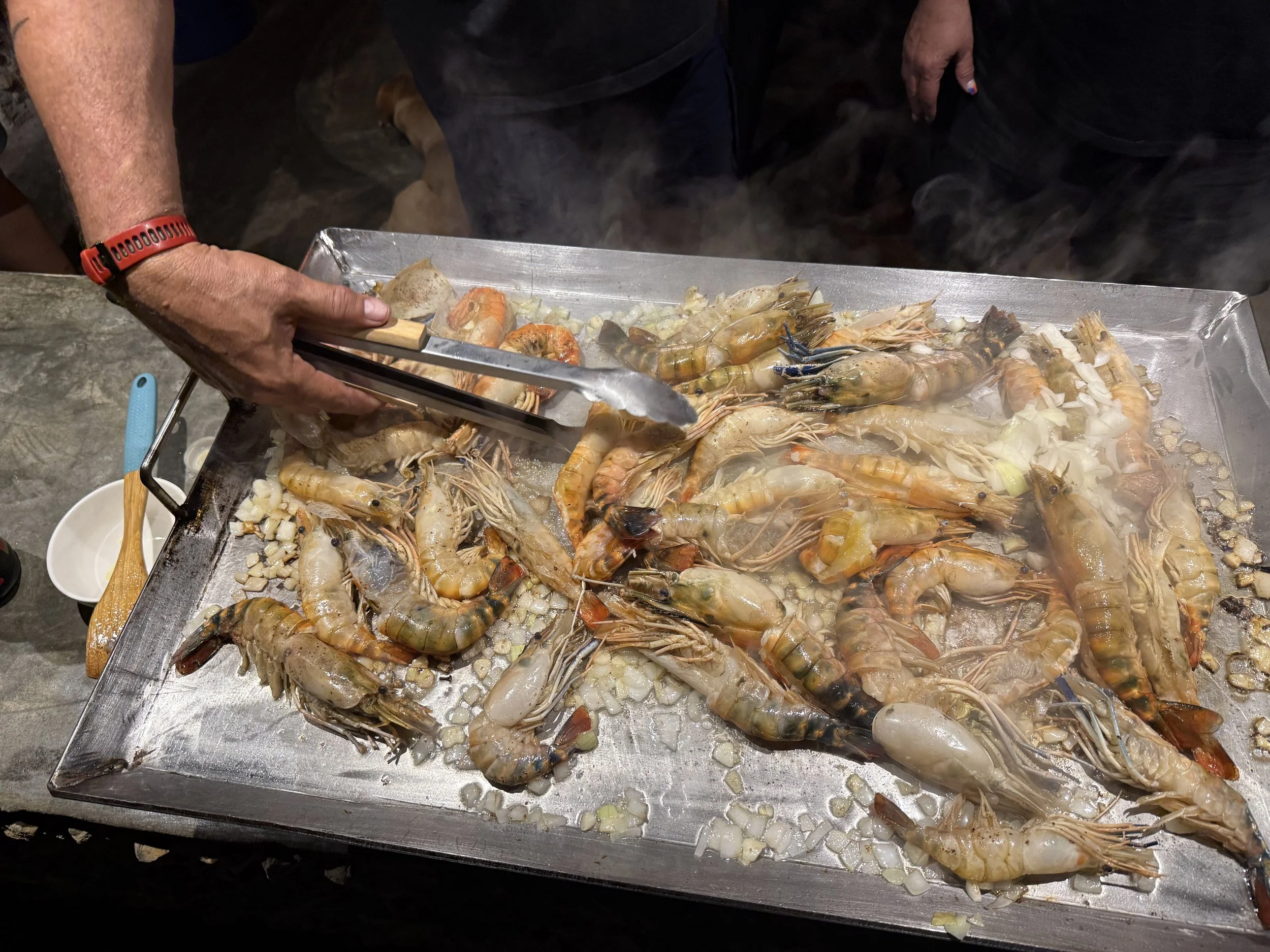 A person preparing a seafood dish with shrimp and other shellfish on a metal tray, using tongs to handle the seafood.