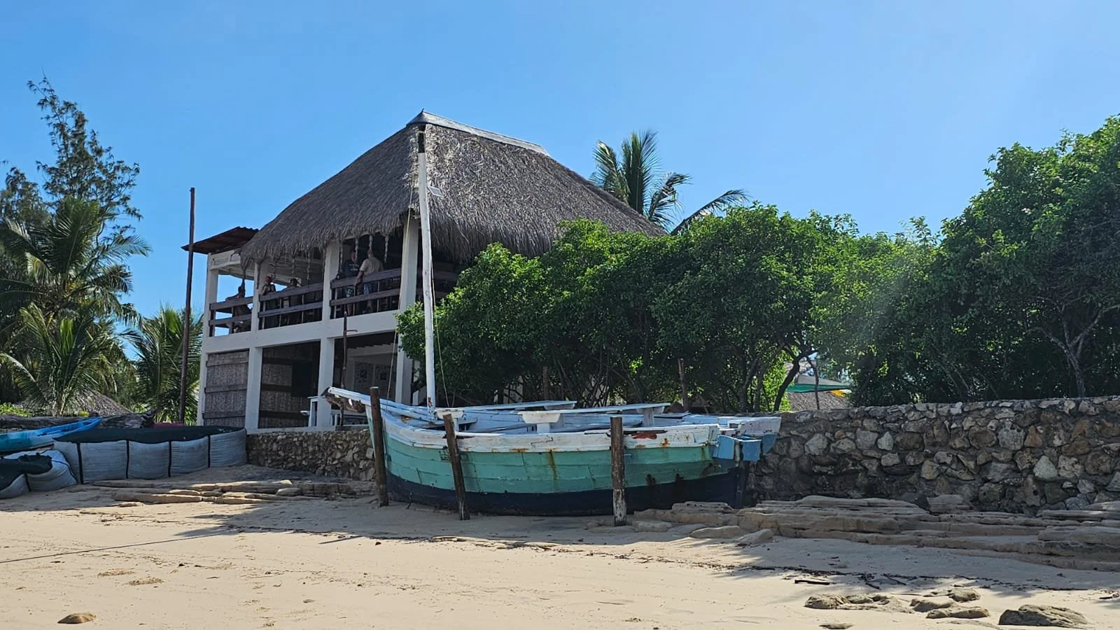 A beachside scene with a thatched-roof hut on stilts, a boat resting on the sand, and lush green trees under a clear blue sky.
