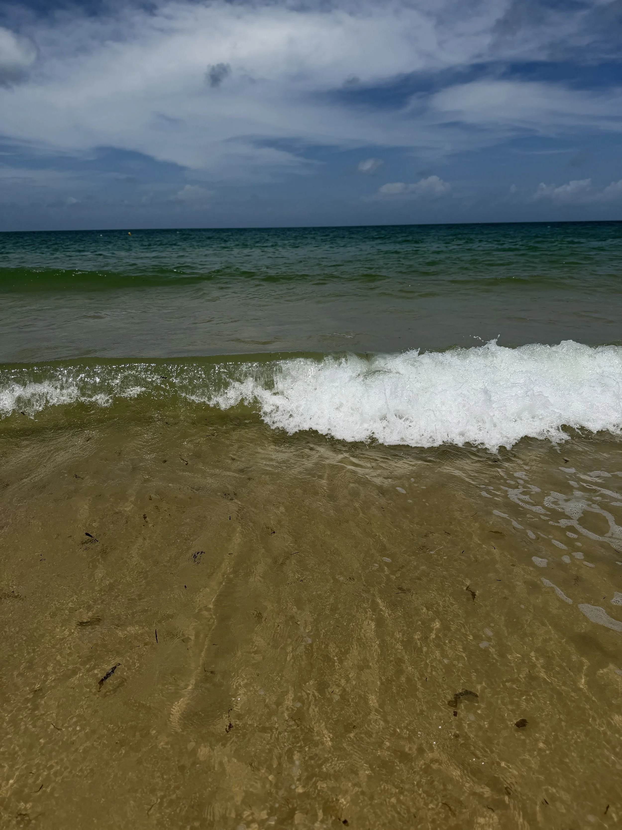 Photo of the ocean with gentle waves hitting the sandy beach under a partly cloudy sky.