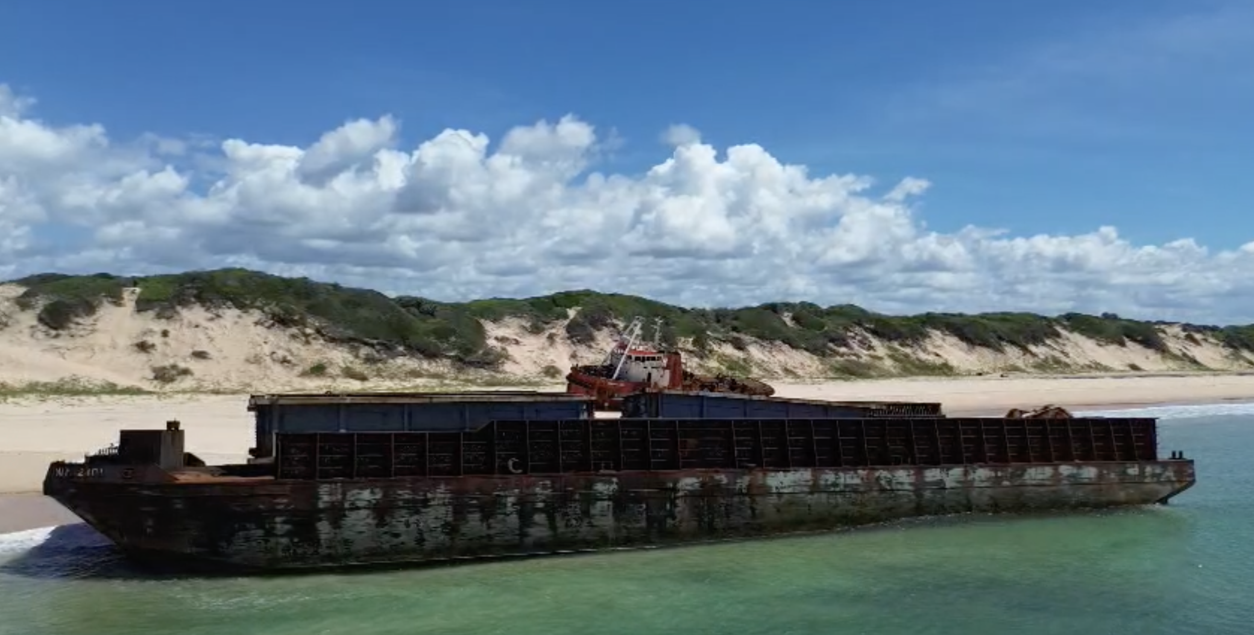 A shipwreck on a sandy beach with green hills and a partly cloudy blue sky in the background.