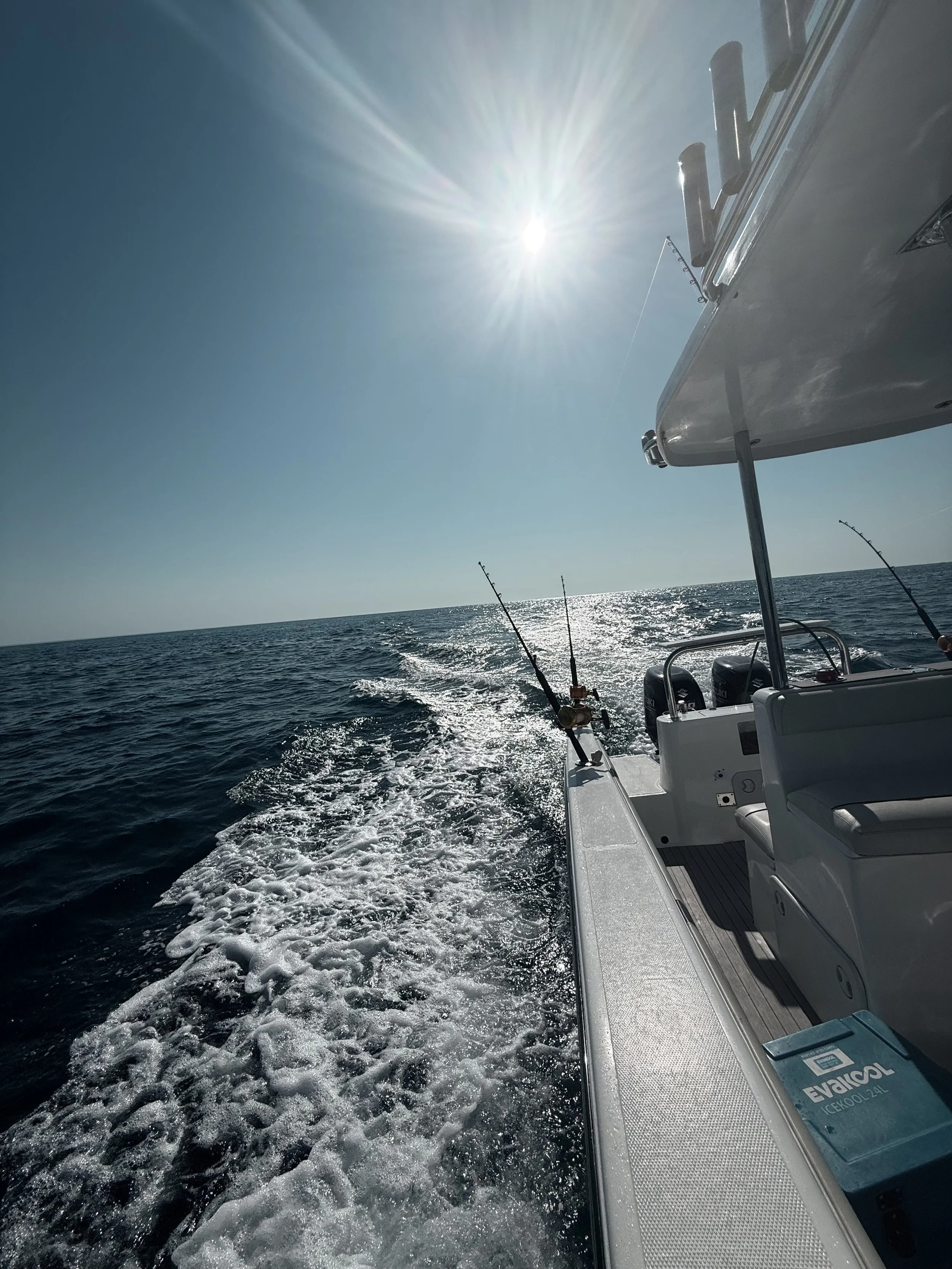 A boat sailing on the ocean with fishing rods mounted on the side, under a bright sun with rays shining down, creating a glare on the water.