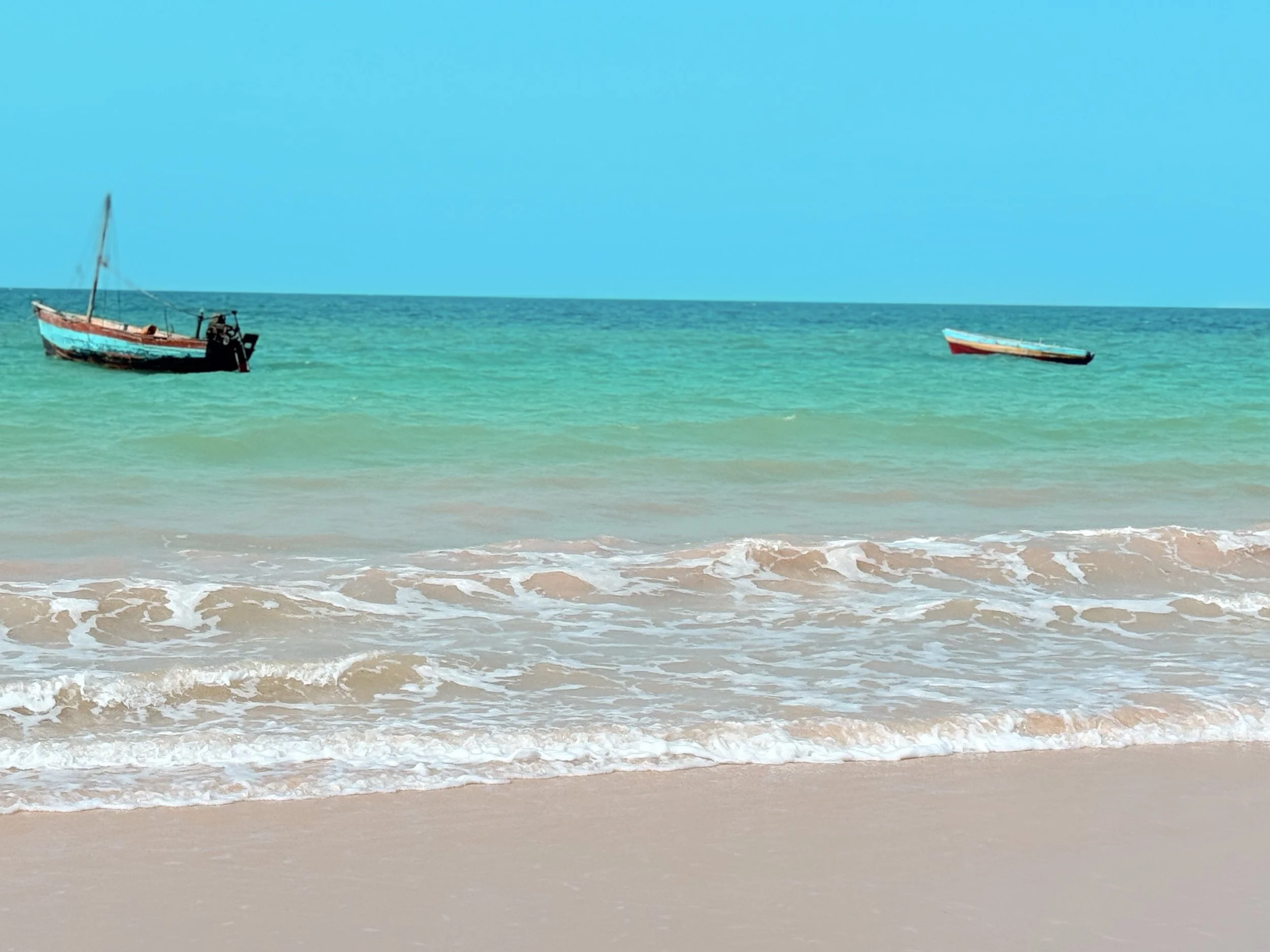 View of a beach with waves crashing on the shore, two small boats floating on the water, and a bright blue sky.