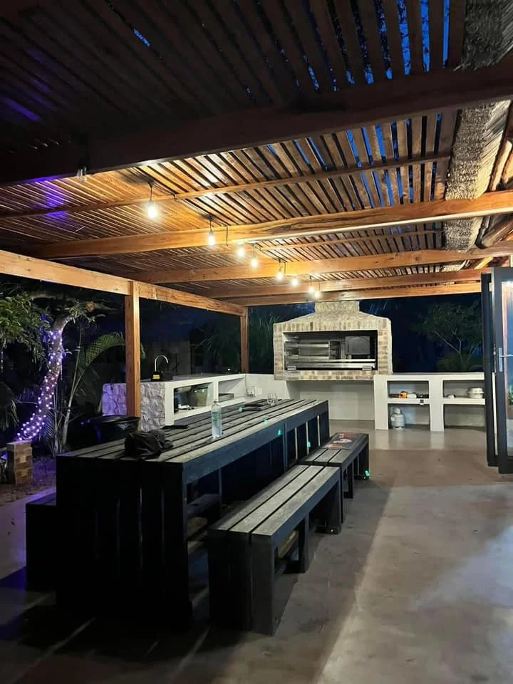 Outdoor patio area with a wooden slatted roof, string lights, and a built-in brick barbecue grill. There is a long black wooden table with matching benches and a bottle of water on top. Surrounding the area are trees decorated with purple string lights.