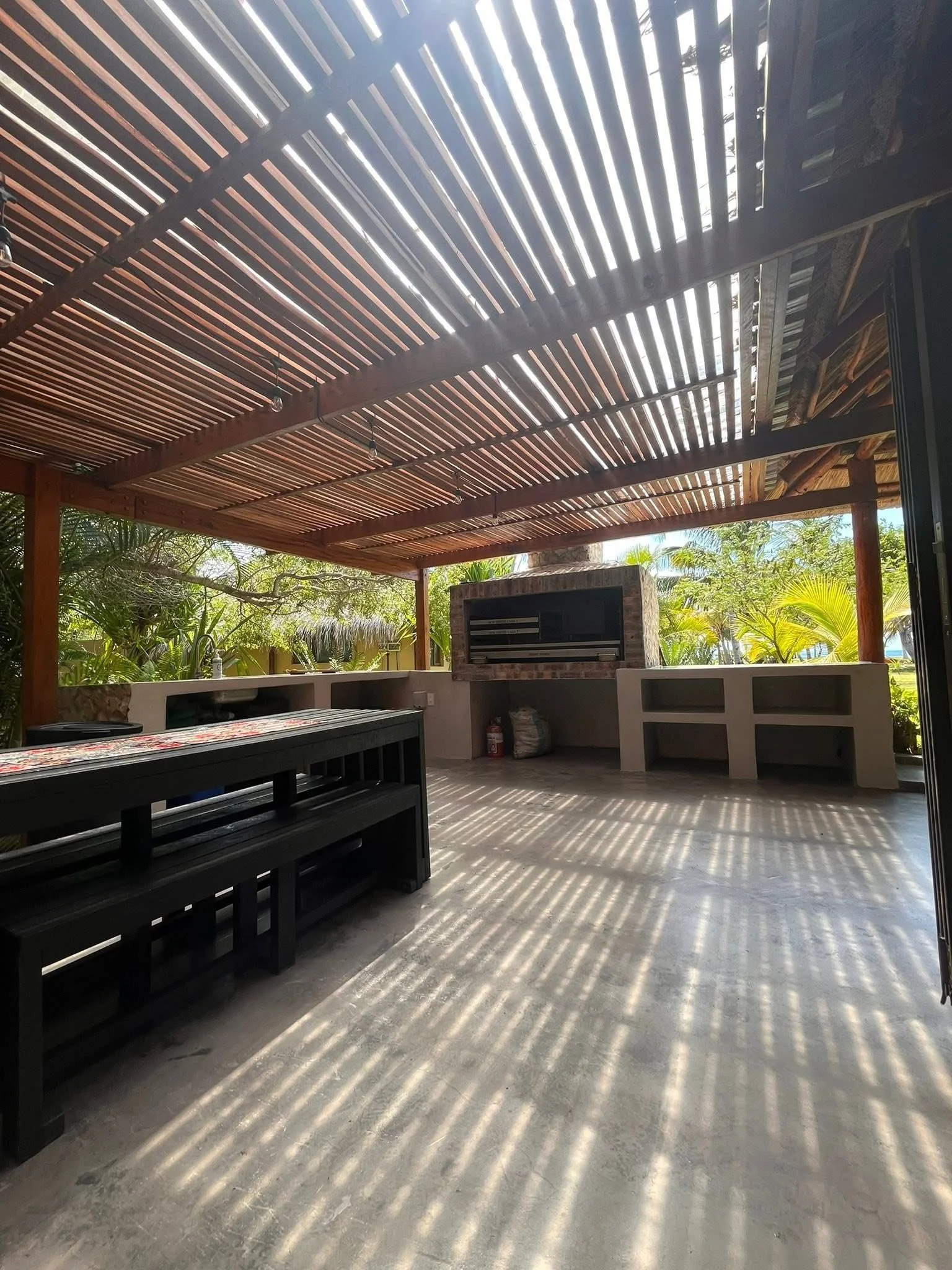 Outdoor patio with a pergola made of wooden slats, casting striped shadows on the concrete floor, with lush green plants and trees in the background.
