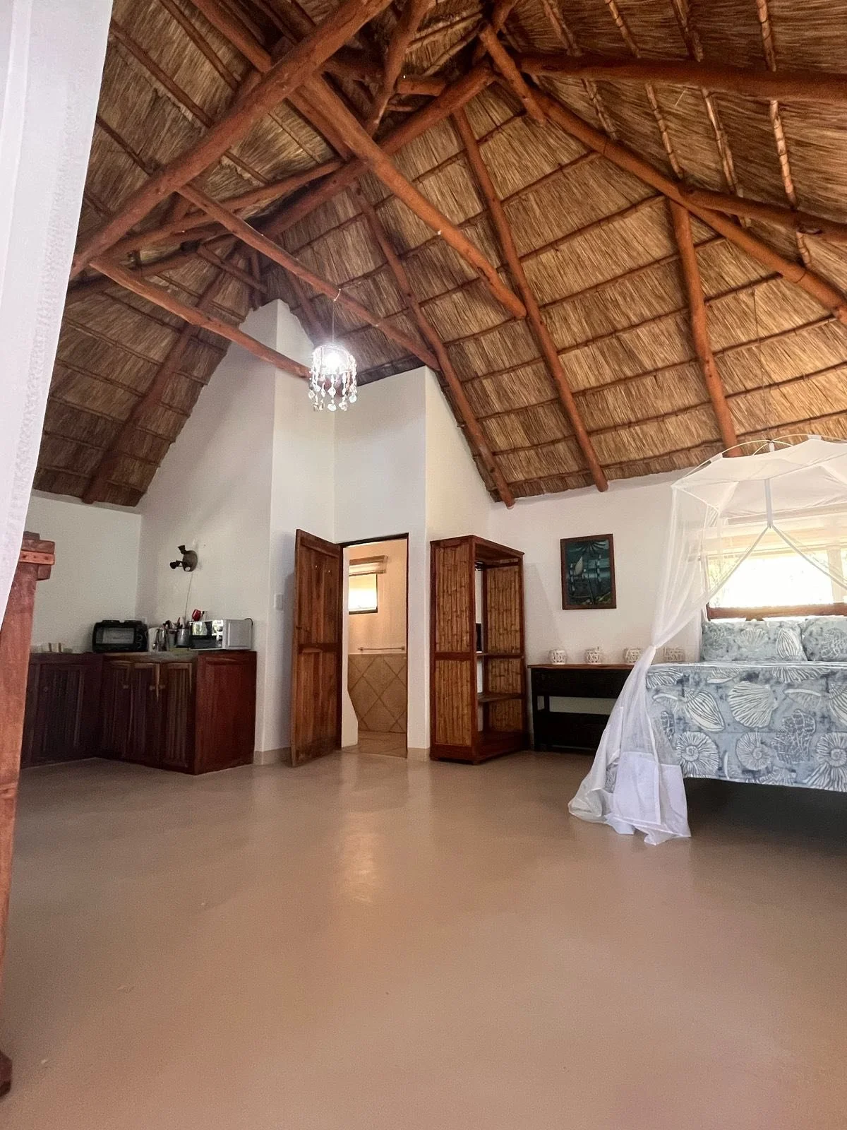 Interior of a room with a thatched roof, a bed with a canopy, wooden furniture, and a small kitchenette area.