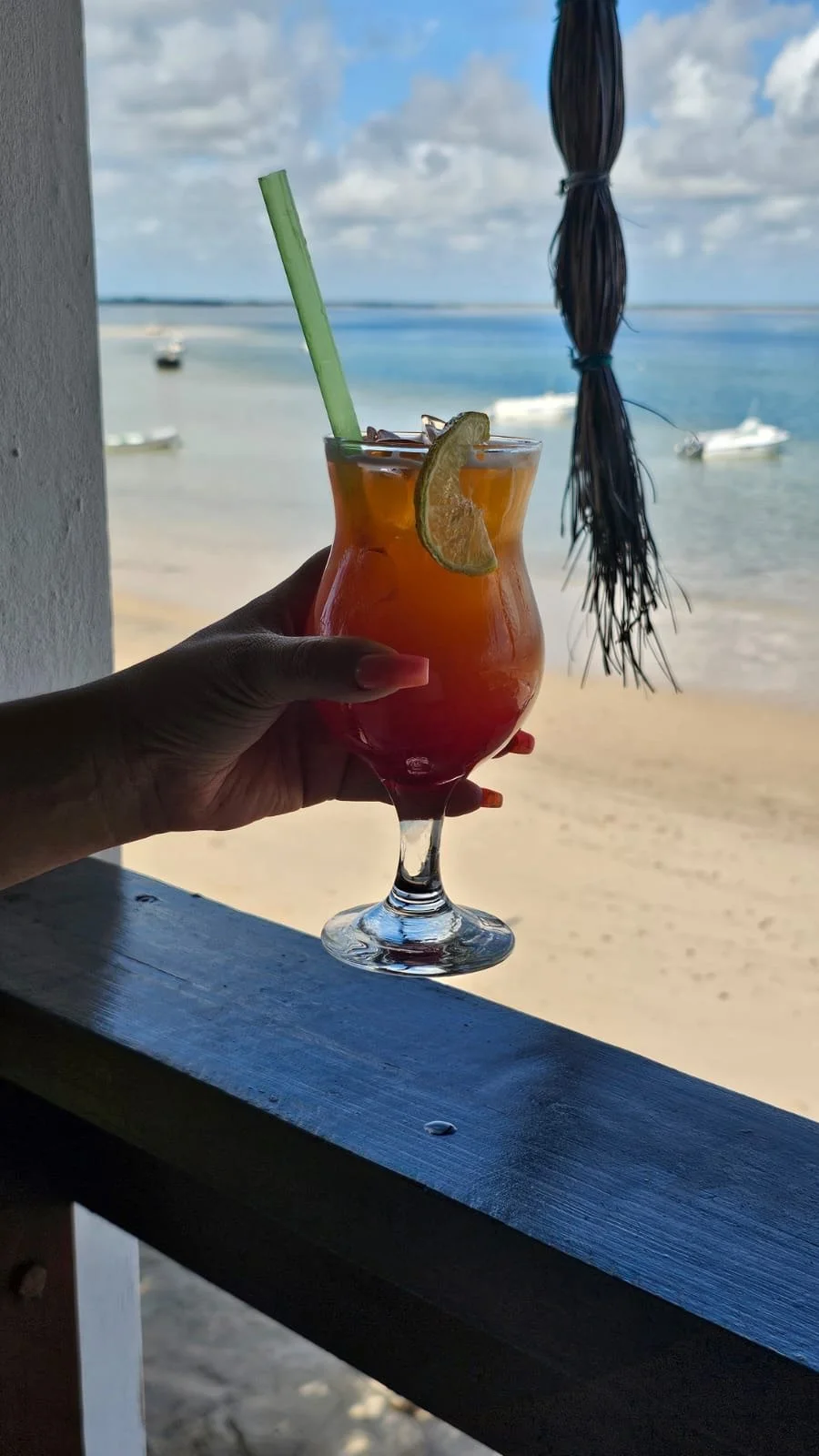 Hand holding a tropical cocktail with an orange slice garnish and a green straw, with a beach and boats in the background.