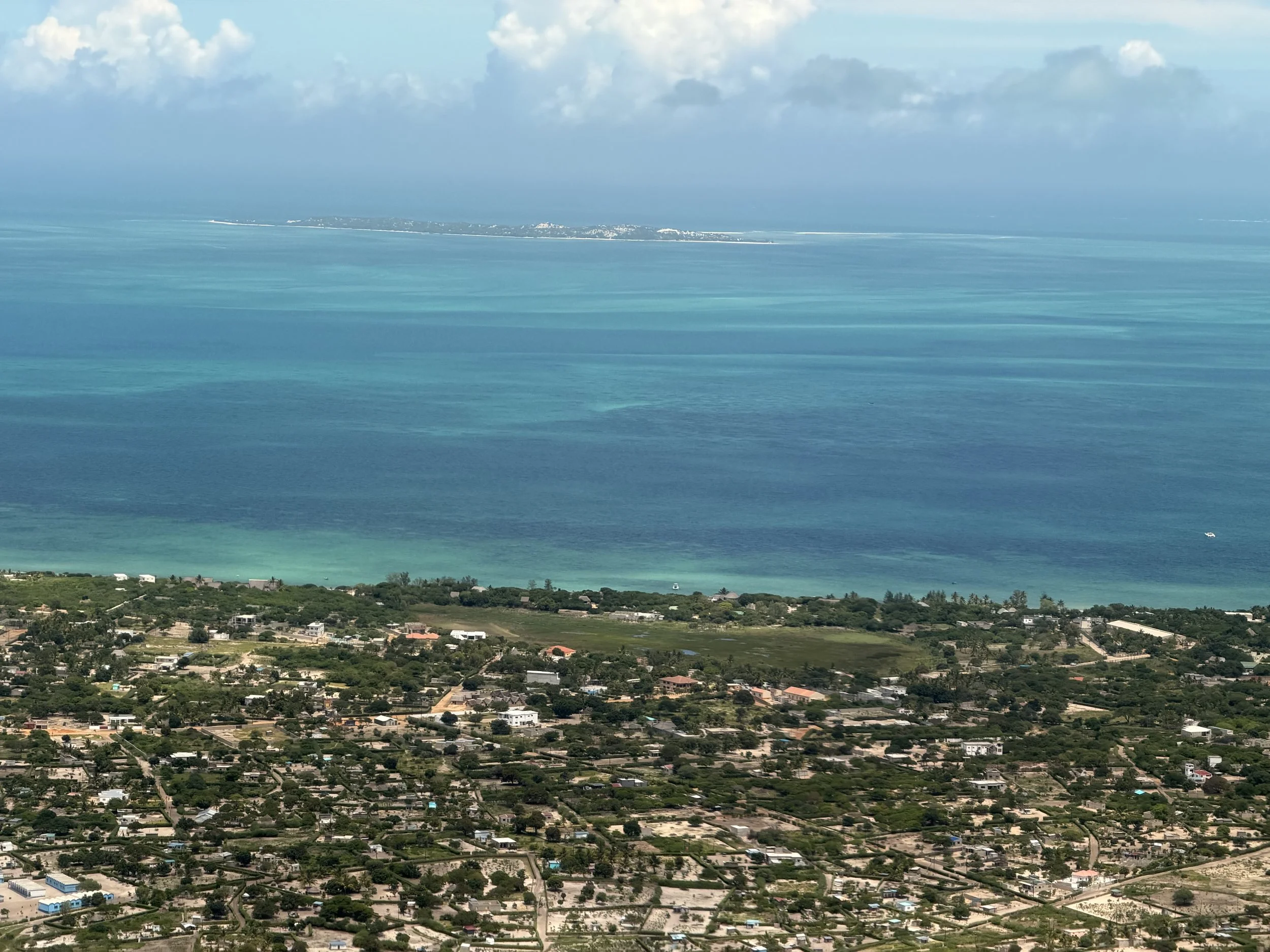 Aerial view of a coastal town with a large body of turquoise water, a sandy beach, and residential buildings with lush greenery.