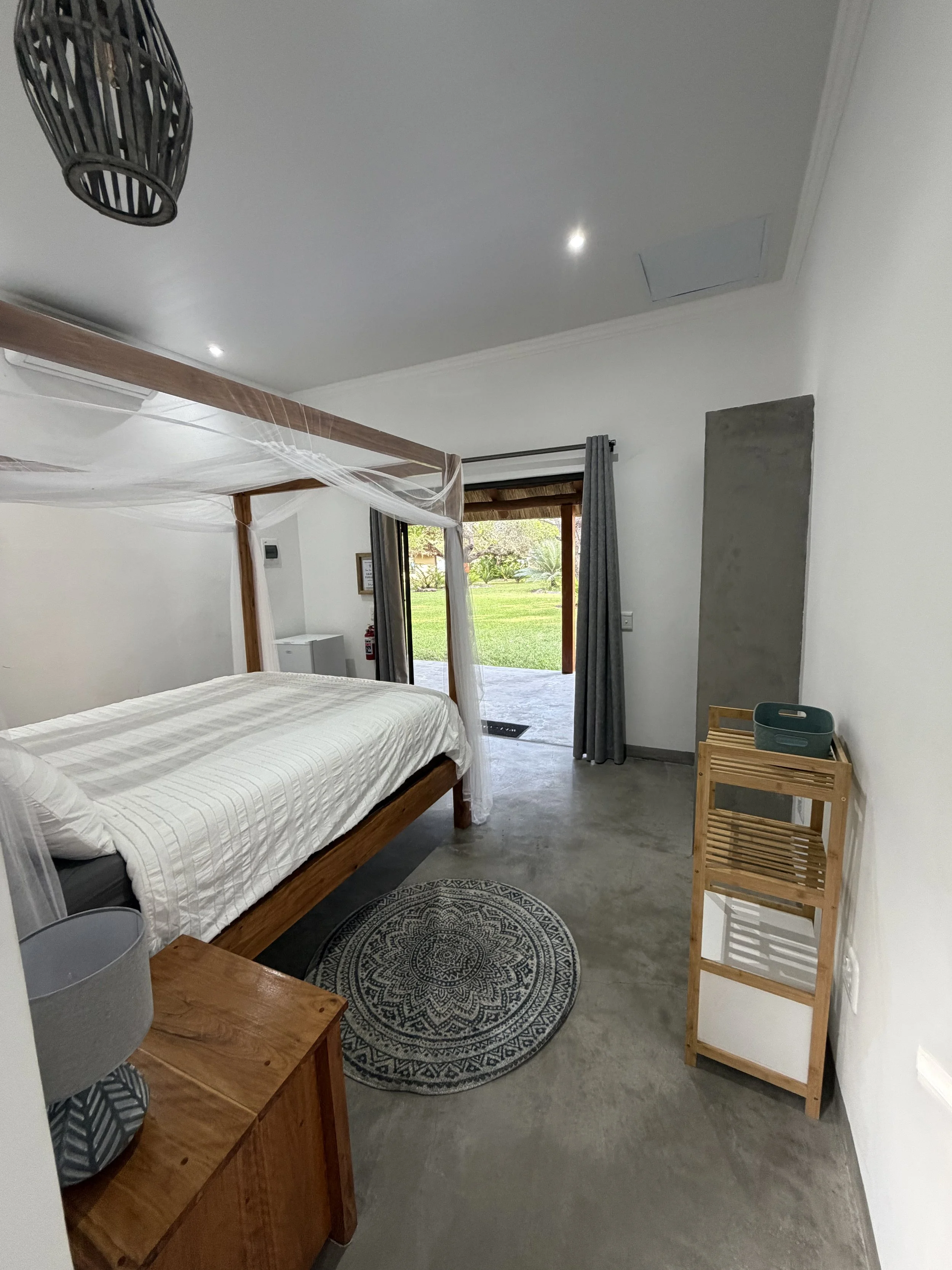 A bedroom featuring a wooden bed with white bedding, a small wooden side table with a gray lampshade, a round patterned rug on the floor, a cylindrical gray basket, and a wooden standing rack with a small basket on top. There is a large sliding glass door with gray curtains that open to a grassy yard with trees outside.