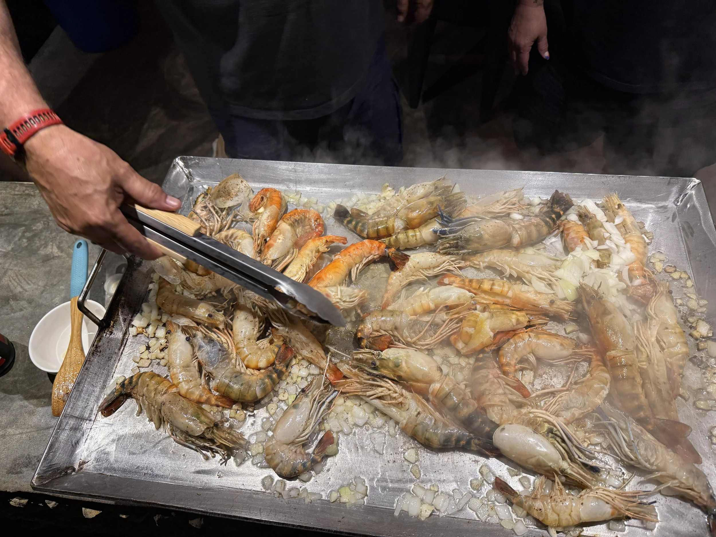 A person preparing a tray of raw shrimp and chopped vegetables, possibly garlic or onion, on a metal baking sheet.